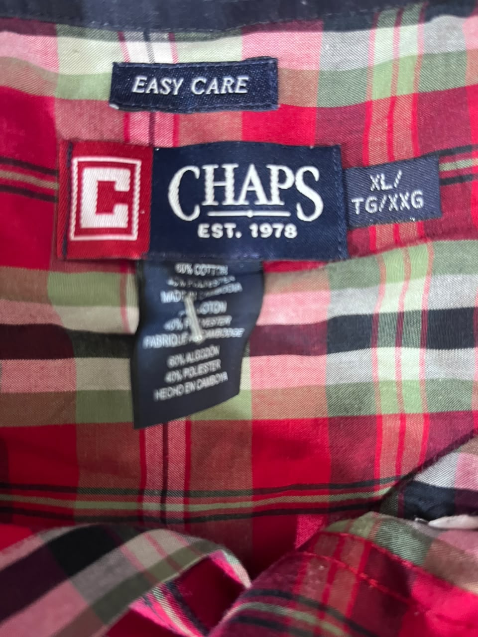 Close-up of a plaid shirt fabric with visible CHAPS brand labels, including an Easy Care label, size label, and care tag. The plaid pattern is primarily red with green, black, and white stripes.