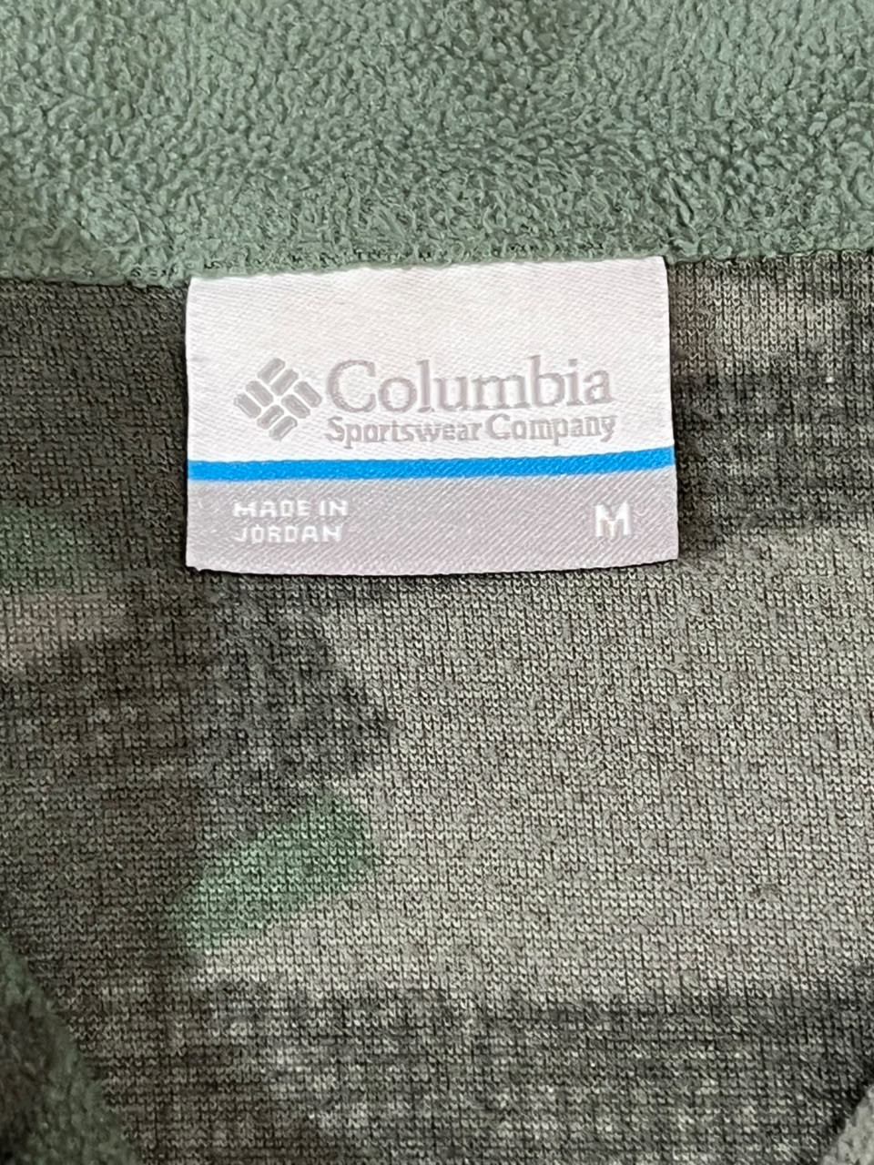 Close-up of a Columbia Sportswear Company size M garment label sewn onto a gray and green textured knit fabric.
