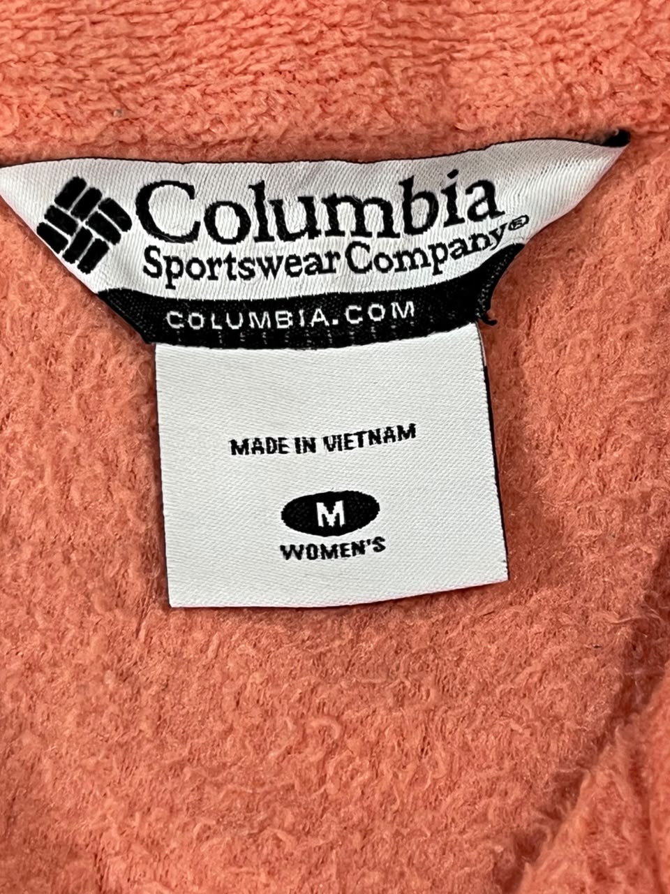 Close-up of a Columbia Sportswear Company garment label on a coral pink textured fabric, showing 'COLUMBIA.COM', 'MADE IN VIETNAM', and 'M WOMEN'S'.