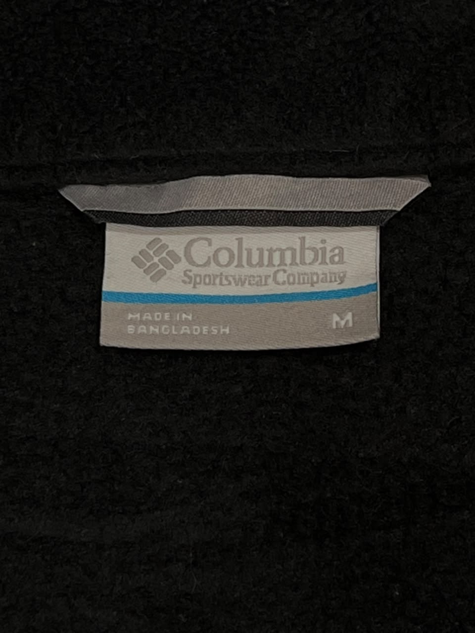 A close-up of a Columbia Sportswear Company fabric tag sewn onto a black garment, showing the brand name, logo, 'Made in Bangladesh', and size 'M'.