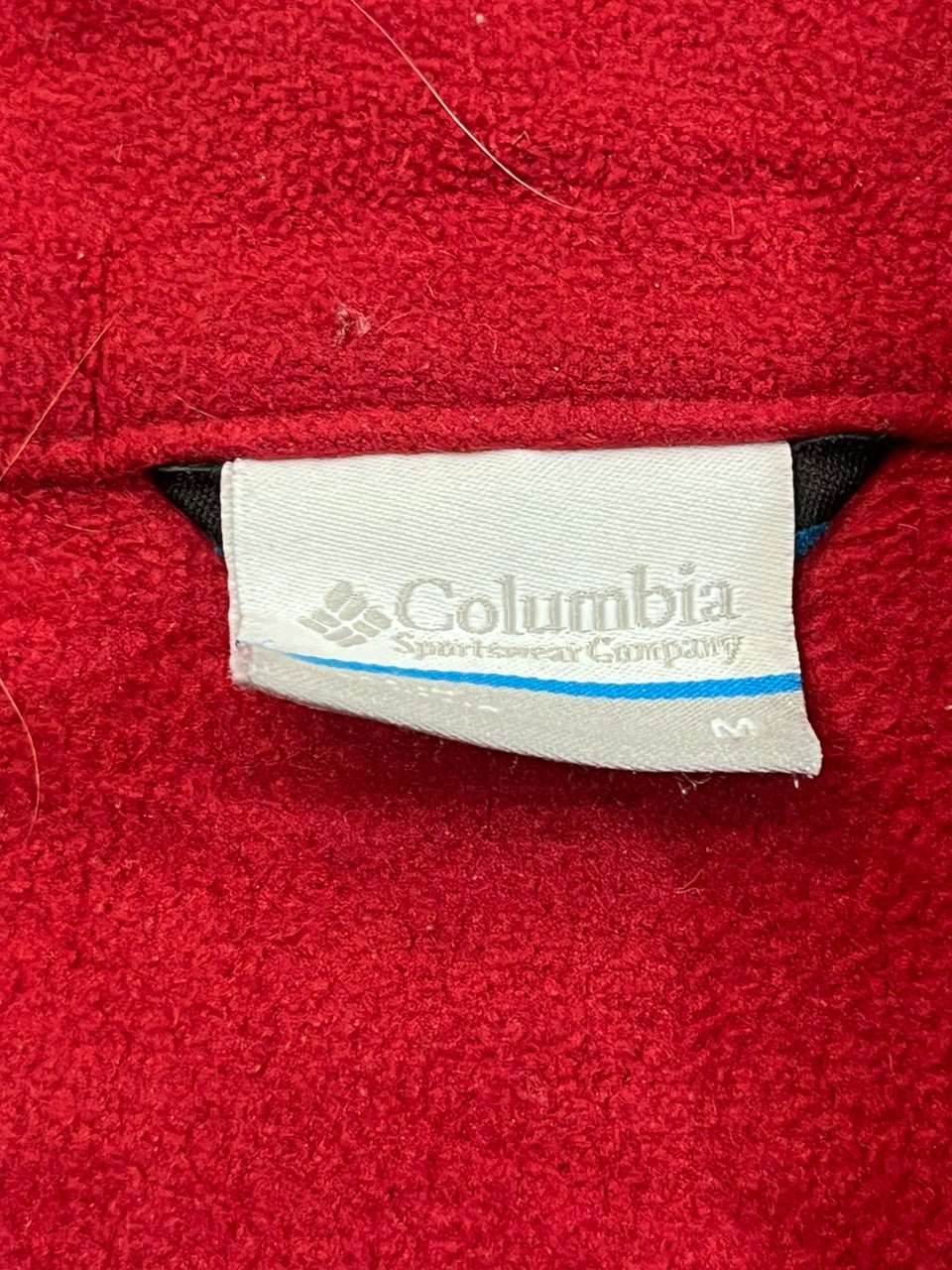 A close-up of a red fleece garment showing the interior neck label, which is white and printed with the Columbia Sportswear Company logo.