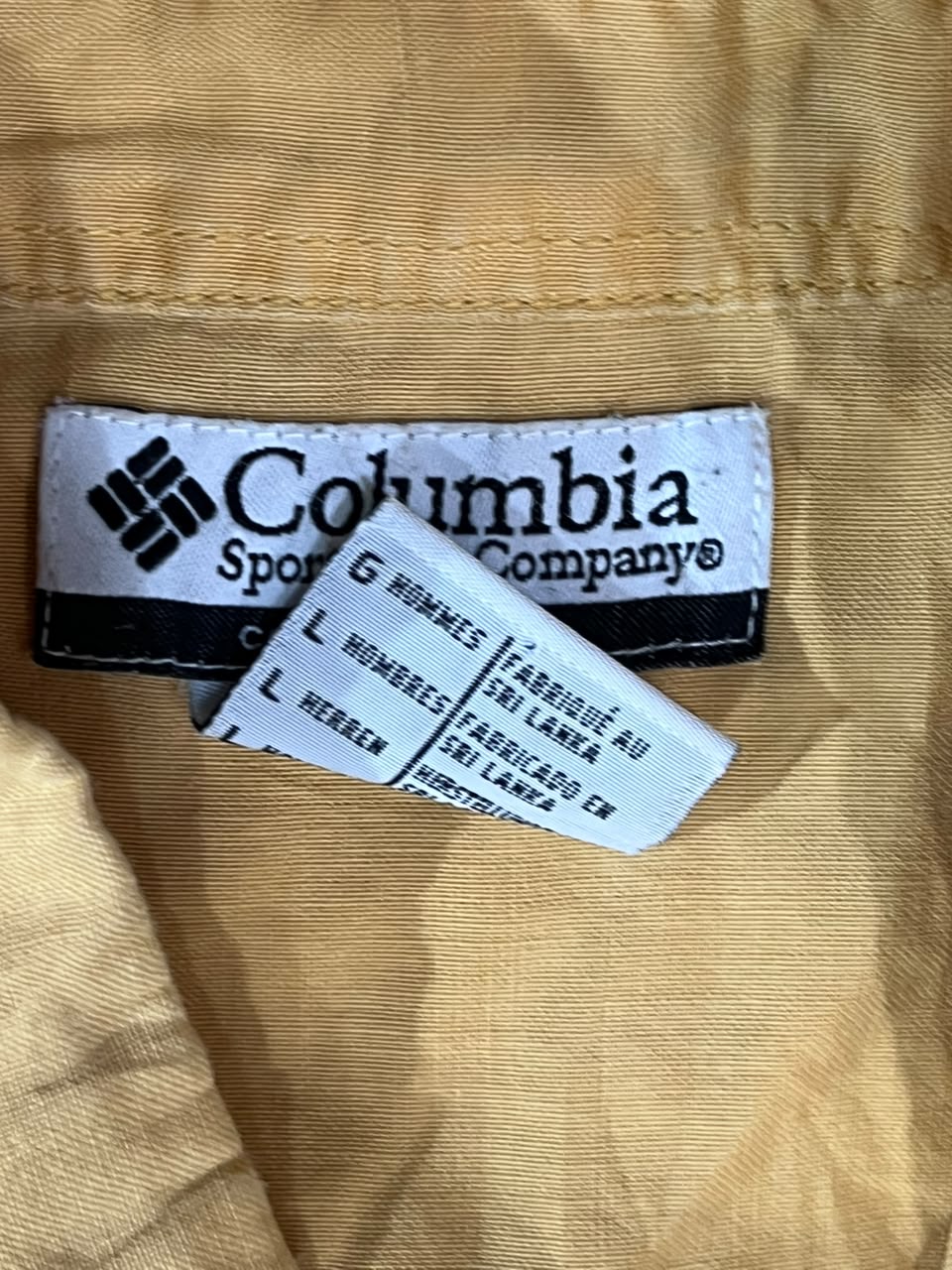 Close-up of a Columbia Sportswear garment label sewn onto tan fabric, with a second care tag partially visible in front.