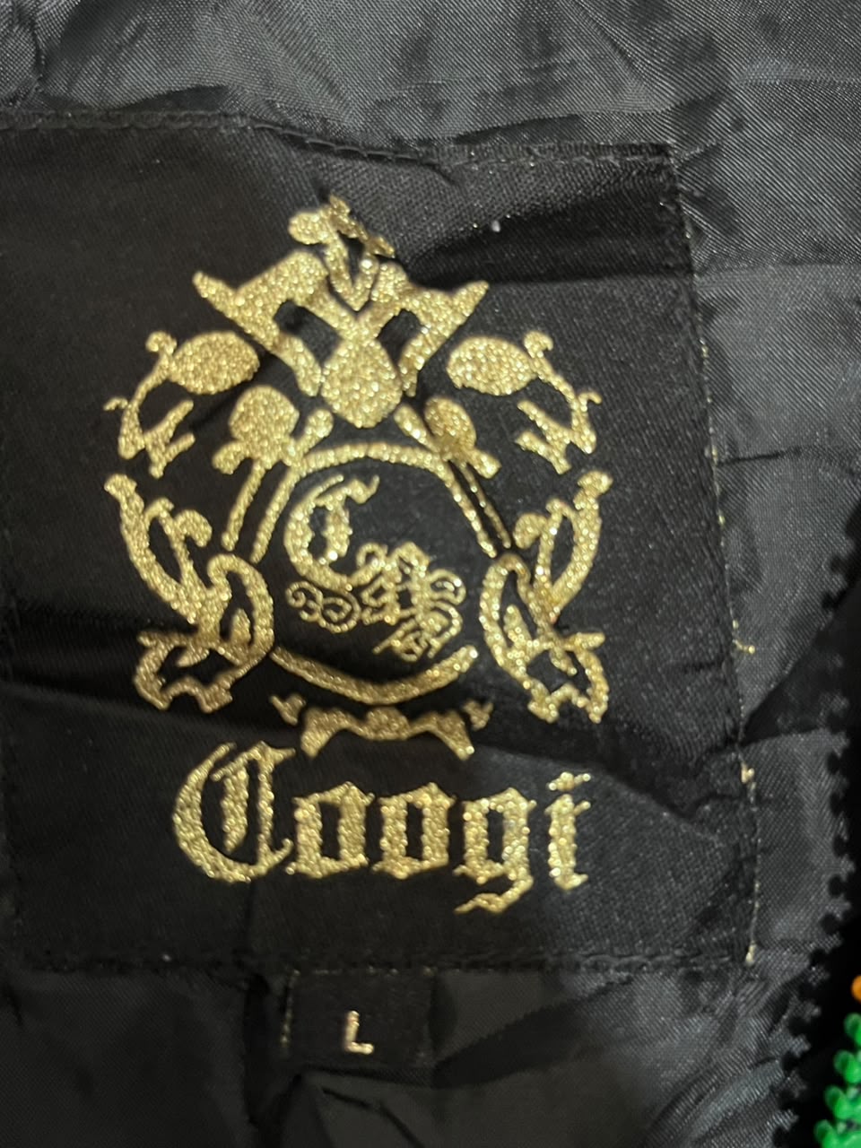 Close-up of a black garment interior label featuring a gold embroidered crest and the word 'Coogi', with a small size tag marked 'L' below.