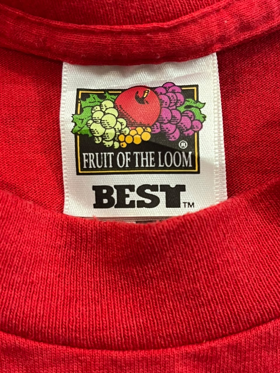 Close-up of a red knit garment showing the inner neck label, which reads 'FRUIT OF THE LOOM' and 'BEST' with a colorful fruit graphic.