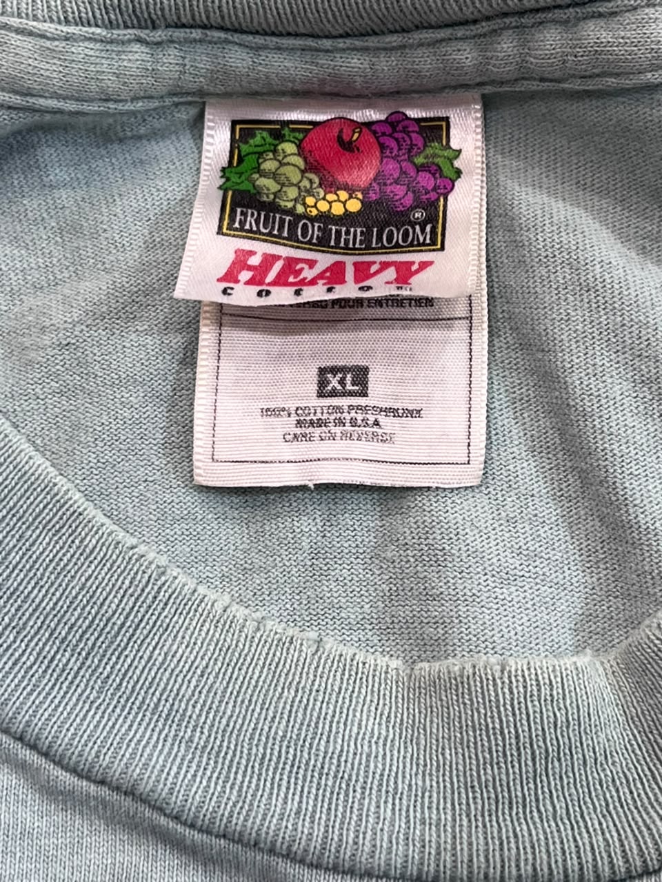 Close-up of a light blue-gray ribbed fabric garment showing the inside neck label, which reads Fruit of the Loom Heavy and shows an XL size tag.