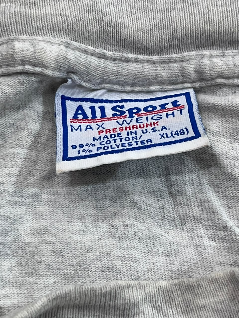 Close-up of a gray heathered garment label reading 'All Sport' with details including 'MAX WEIGHT', 'PRESHRUNK', 'MADE IN U.S.