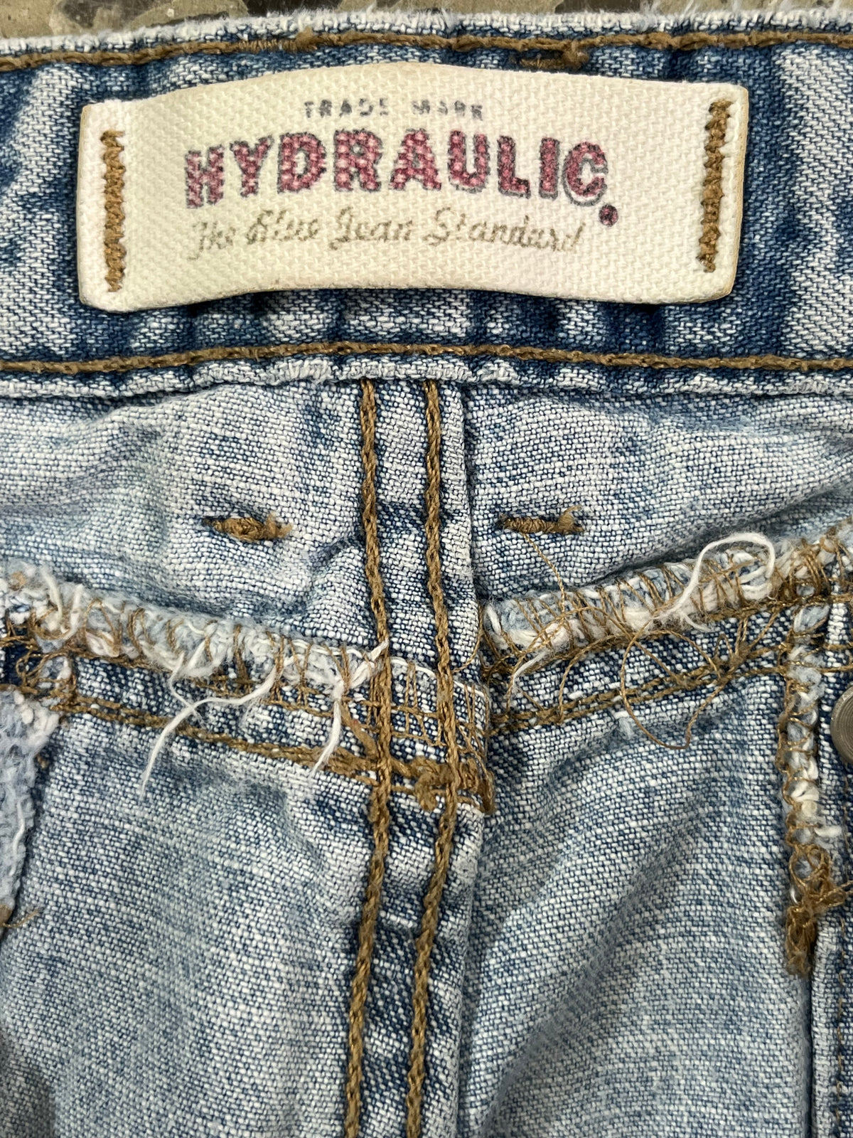 Close-up of the waistband of blue denim jeans showing a sewn-on brand label that reads 'HYDRAULIC The Blue Jean Standard' and detailed frayed stitching.