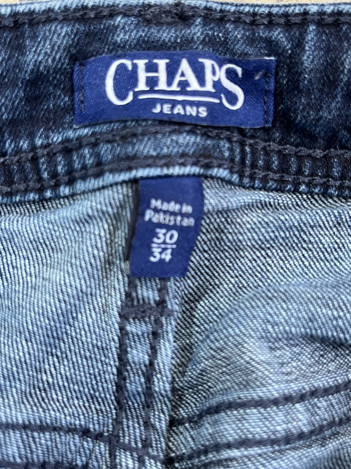 Close-up of a pair of blue denim jeans showing the waistband, with a visible CHAPS JEANS label and a smaller blue size tag reading Made in Pakistan 30 34.