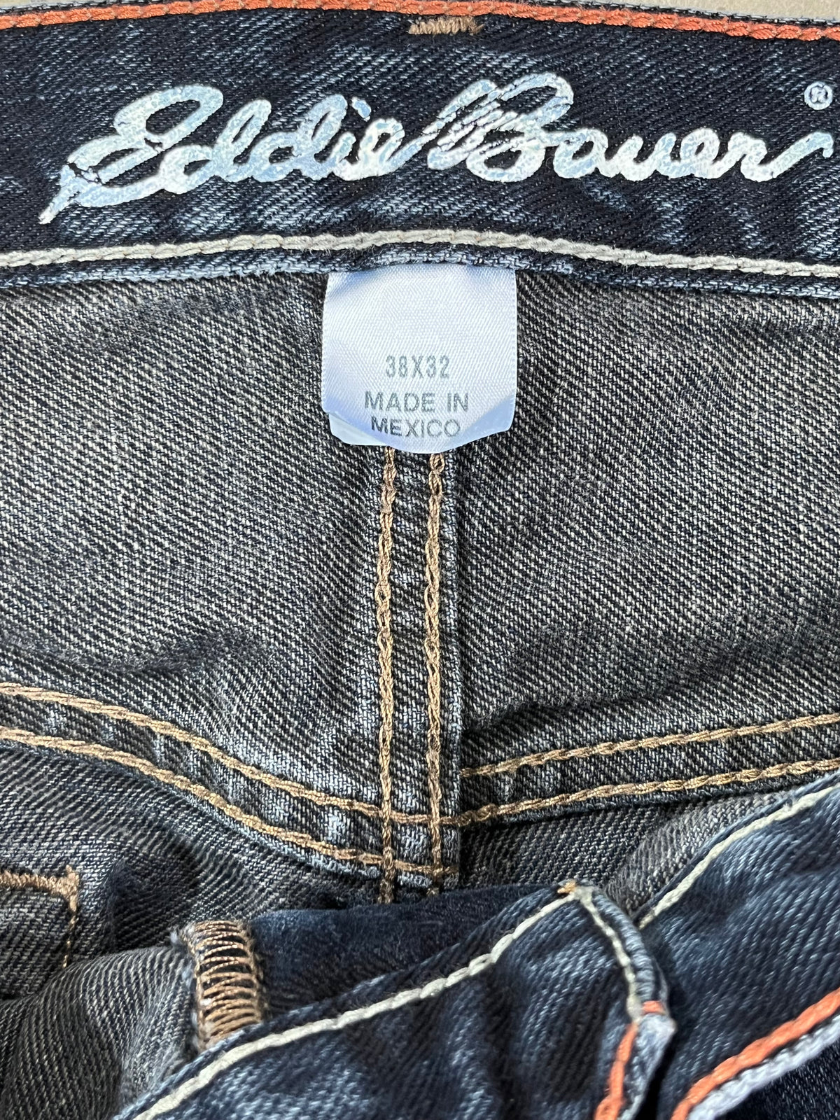 Close-up of a pair of dark-wash denim jeans showing the inner waistband with white embroidered 'Eddie Bauer' branding, a central seam, and a white size/origin label reading '38X32 MADE IN MEXICO'.