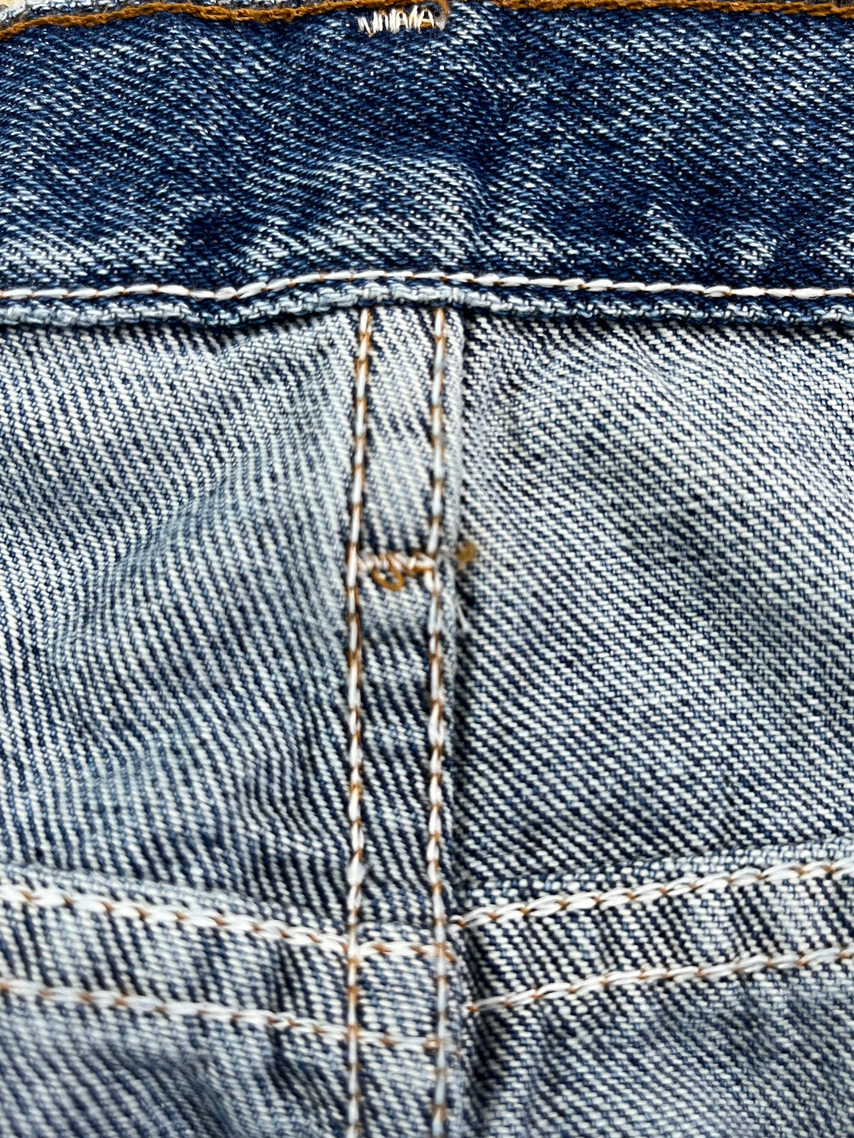 Close-up of a pair of blue denim jeans showing the back pocket construction, including diagonal twill weave, contrast stitching, and a yellow-orange rivet.