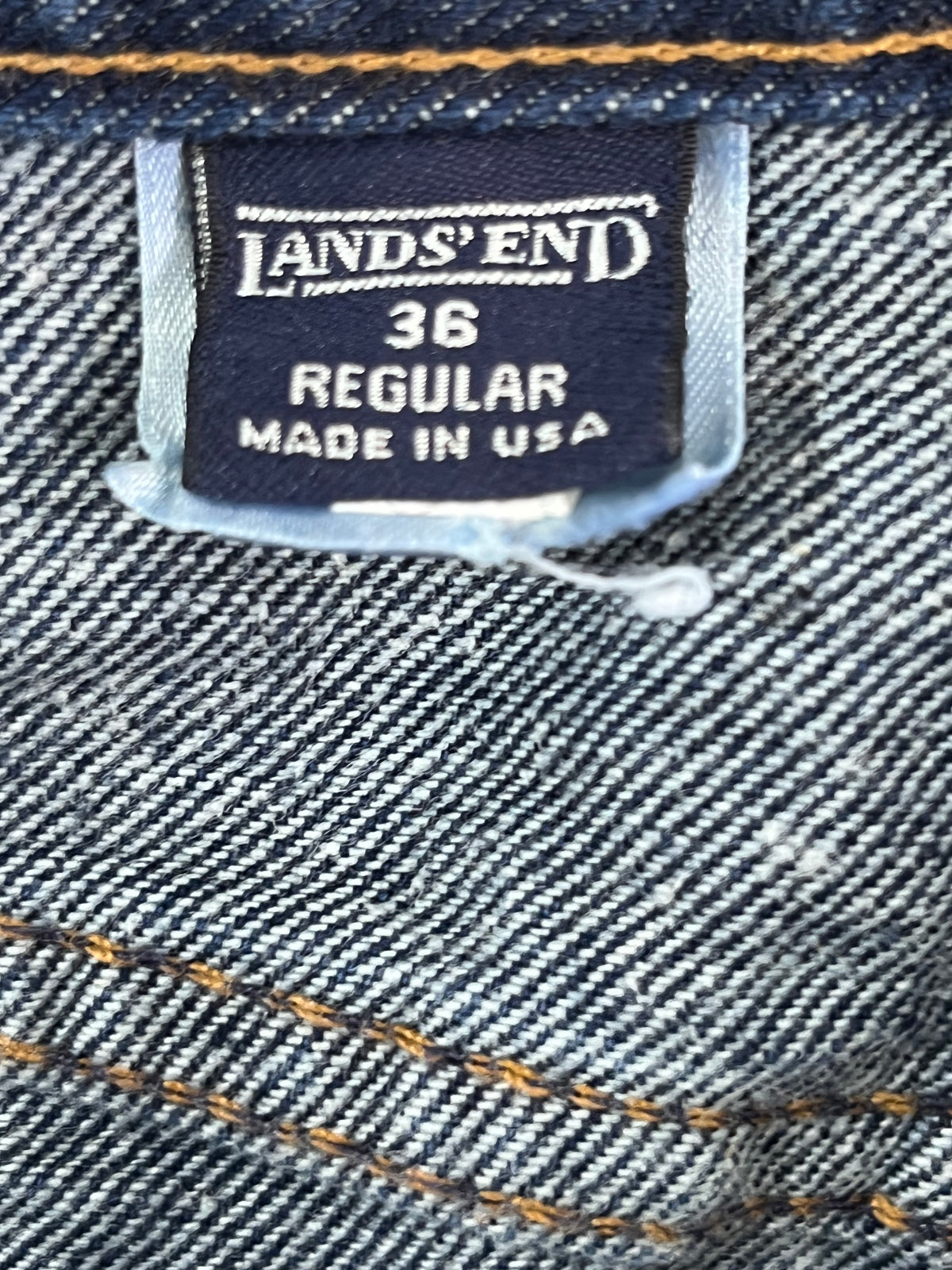 Close-up of a denim garment label reading LAND'S END 36 REGULAR MADE IN USA, with blue-gray twill fabric and orange contrast stitching visible.