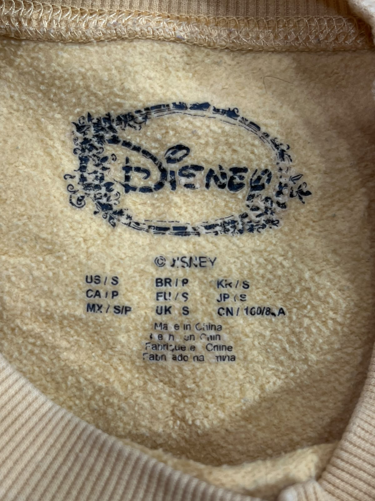 Close-up of the inside collar of a beige knit garment showing a dark blue printed Disney logo and size information on the fleece lining.