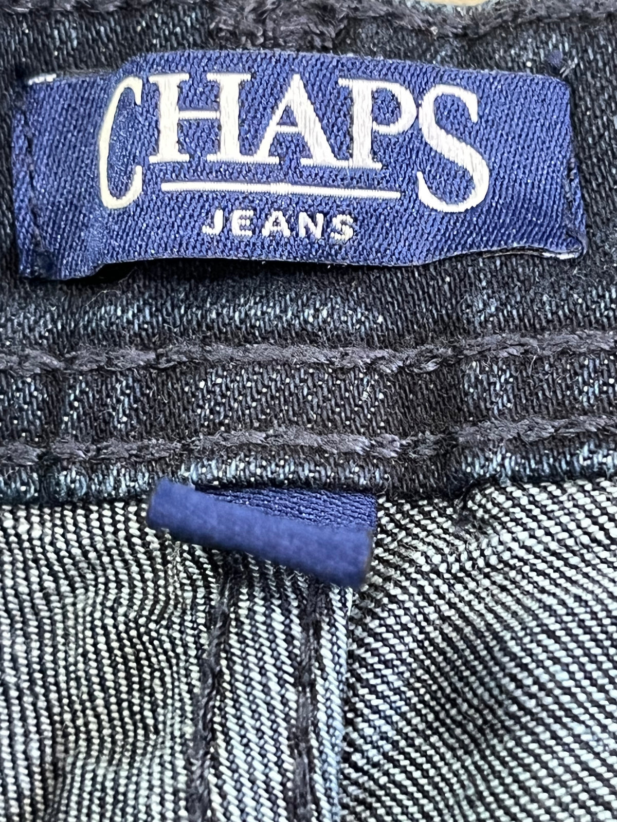 Close-up of a denim waistband showing a blue woven label that reads CHAPS JEANS in white text, with the blue brand tab and striped denim fabric visible.