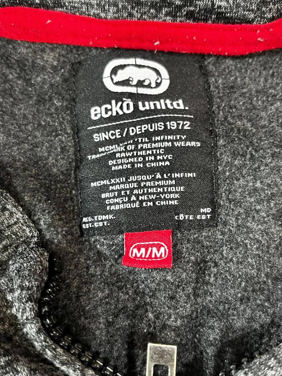 Close-up of a dark gray knit garment label reading 'eckö unidt.' and 'SINCE / DEPUIS 1972' with a red 'm/m' tag below it, plus a red trim line at the collar.