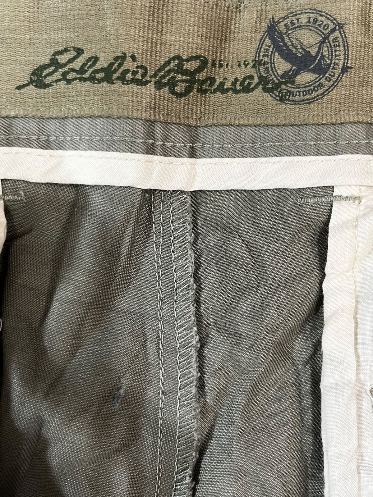 Close-up of a vintage pair of Eddie Bauer shorts showing the interior waistband with the brand's signature script logo and circular bird emblem, alongside gray-green fabric and white trim.