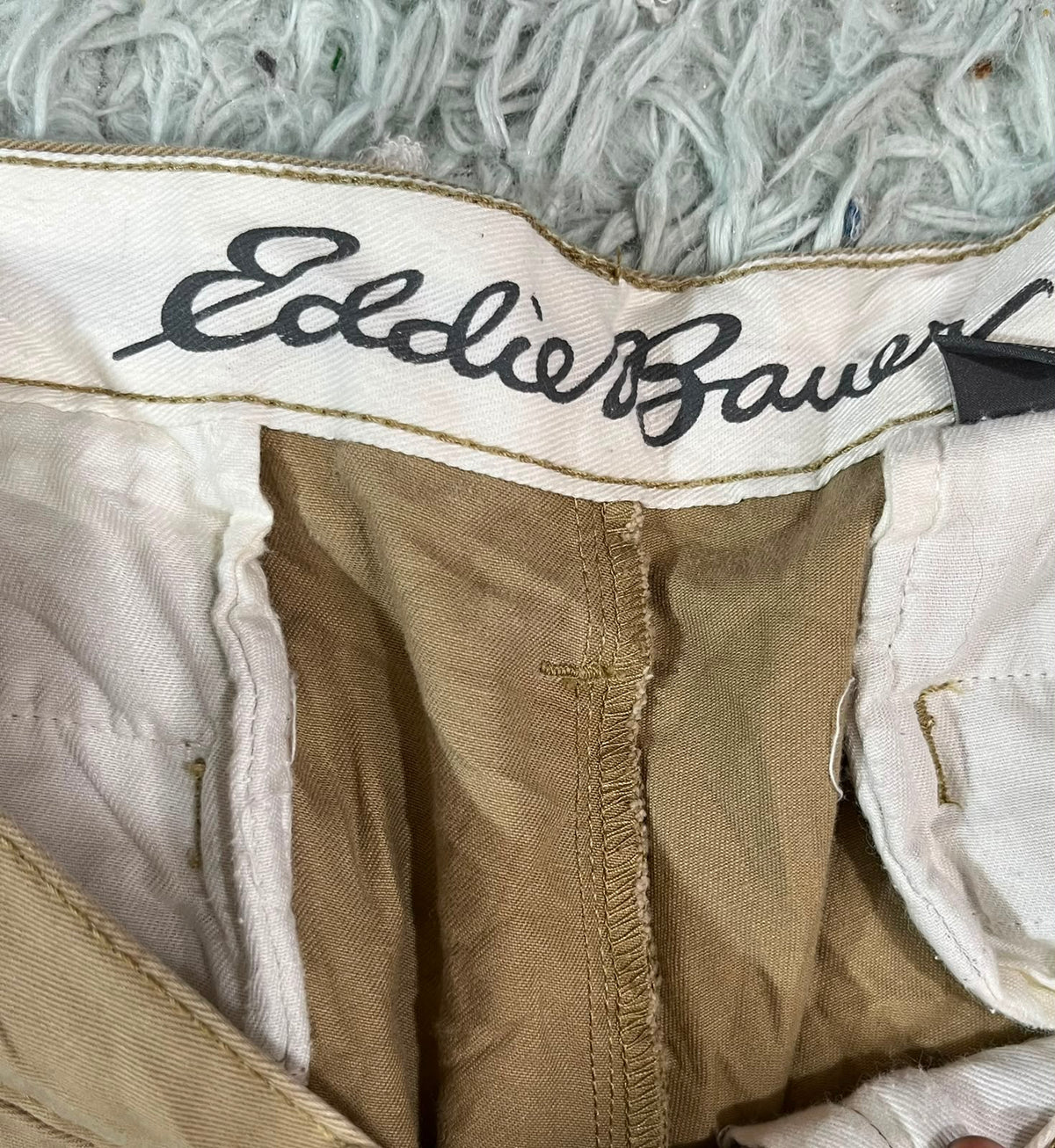 Close-up of a vintage pair of Eddie Bauer shorts showing the interior waistband with the black embroidered brand name on a white canvas band, tan fabric lining, and a light gray shag rug background.