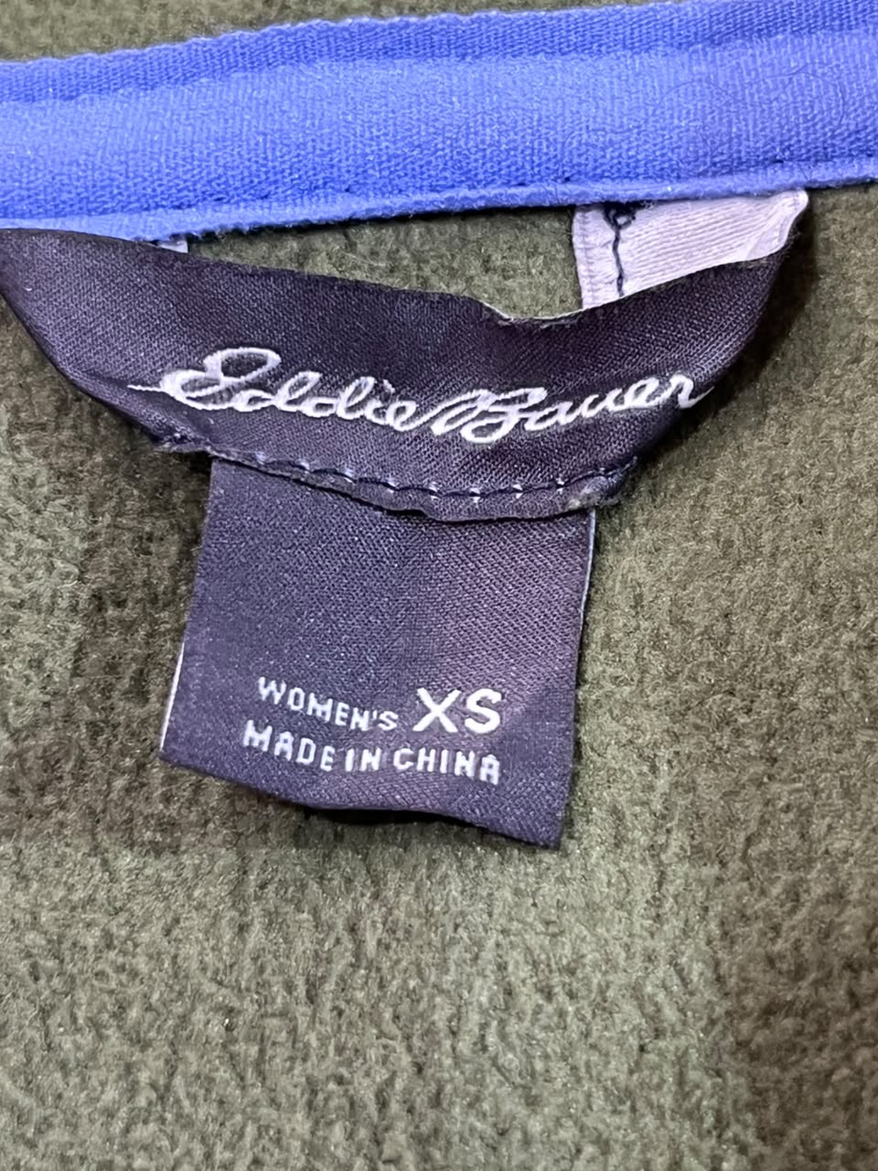 Close-up of a clothing label showing the brand name 'Eddie Guaran' in white script on a dark navy tag, with 'WOMEN'S XS MADE IN CHINA' printed below. The fabric surrounding the label is olive green.