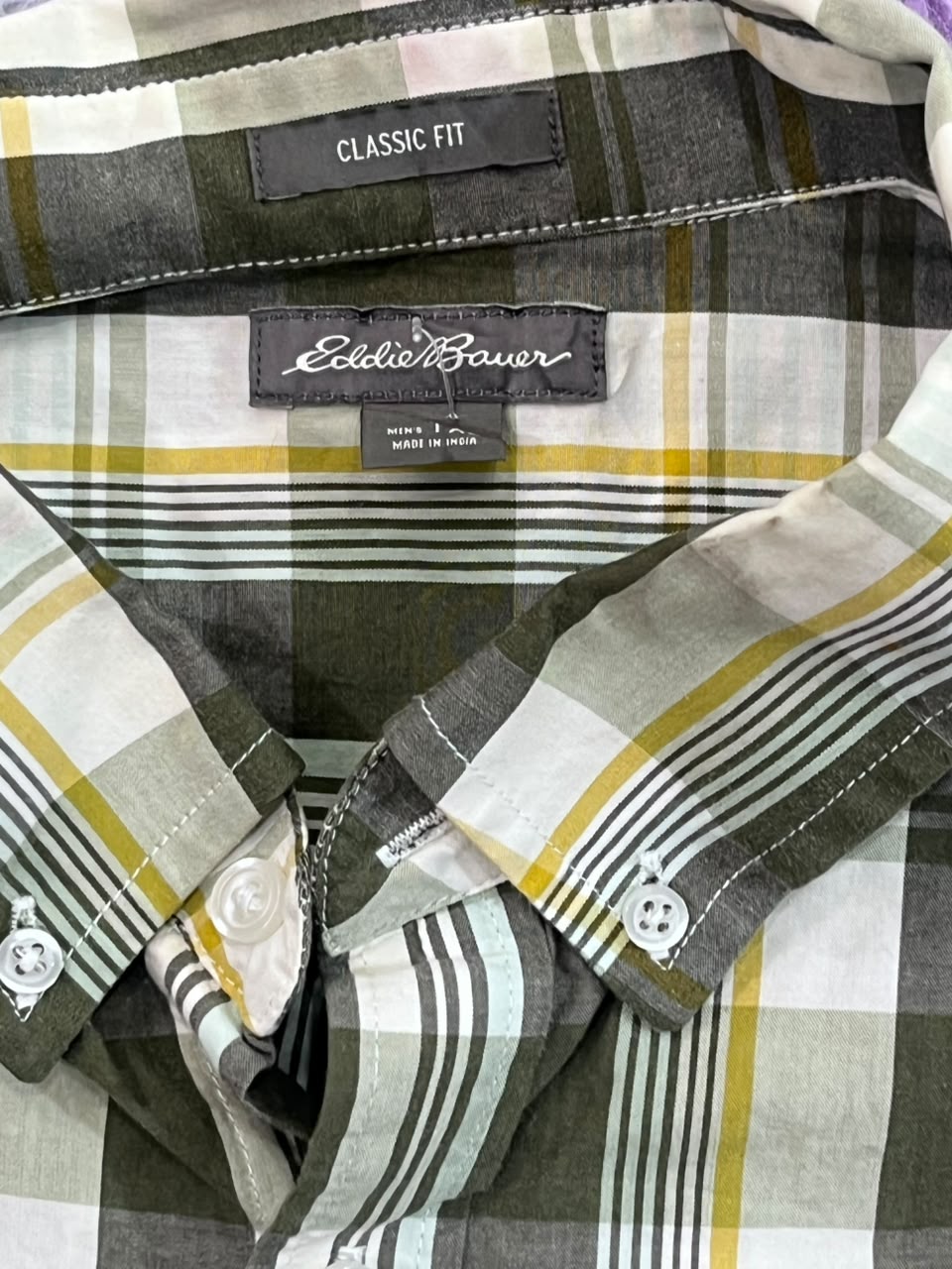 Close-up of an Eddie Bauer plaid button-down shirt collar and inner neck area, showing the brand label, 'CLASSIC FIT' tag, and pattern details in gray, white, yellow, and black.