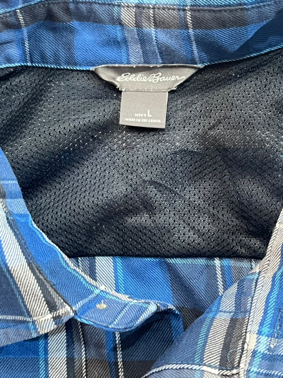 Close-up of the inside collar of a blue plaid button-down shirt with a black mesh lining. The brand tag reads 'Essentials' and the size tag reads 'm/l'.