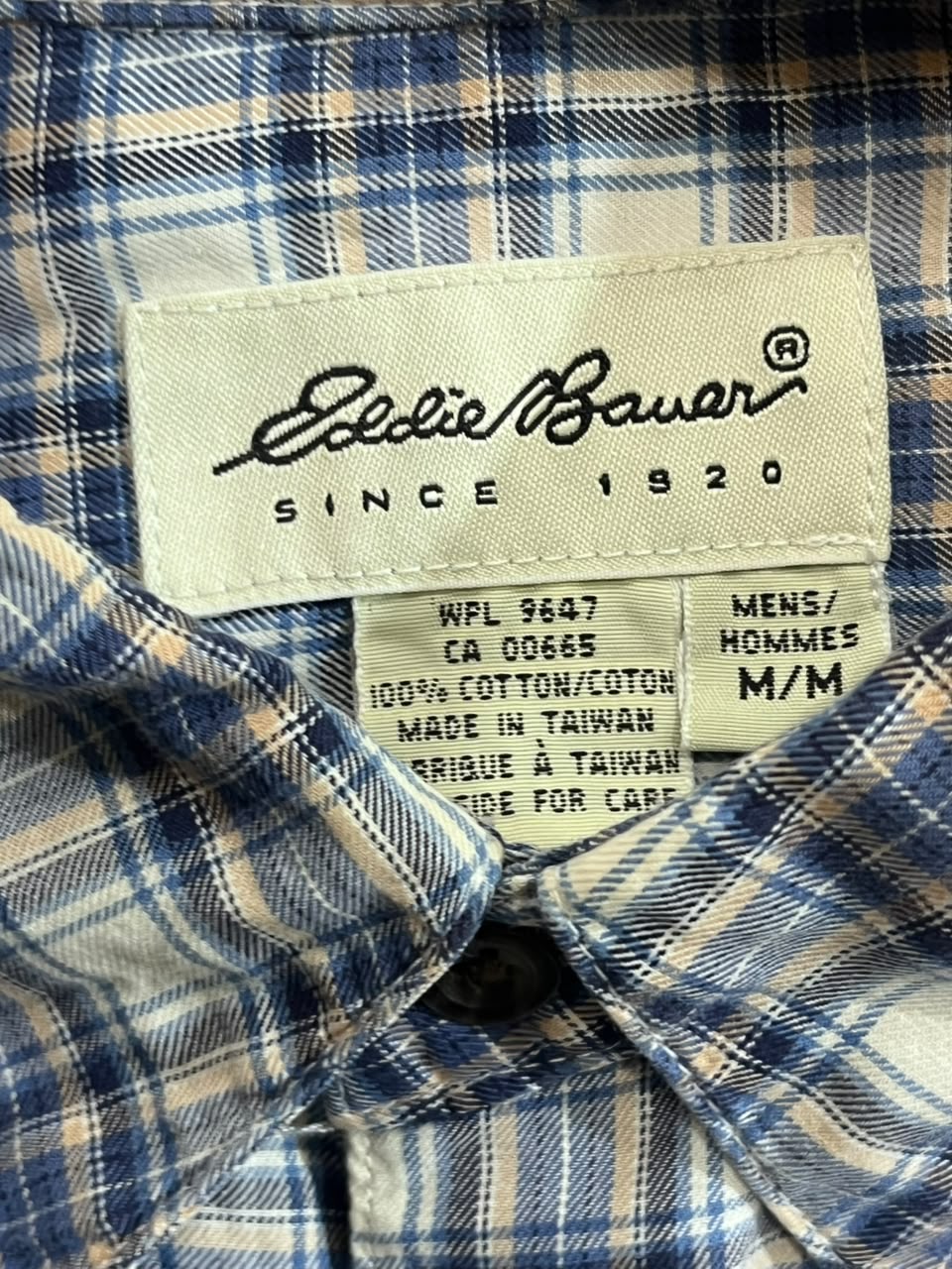 Close-up of a plaid button-down shirt collar with a visible Eddie Bauer neck label. The fabric has a blue, white, and beige plaid pattern, and the label reads 'Eddie Bauer Since 1920' along with care and size information.