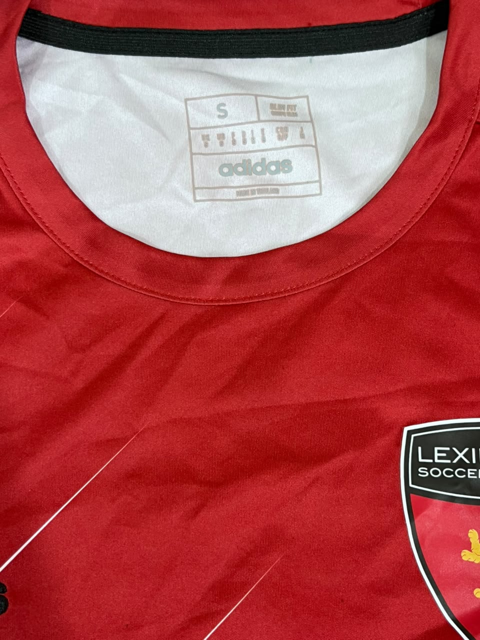Close-up of a red Adidas soccer jersey showing the white inner collar with the Adidas size and brand label, a black trim at the neckline, and part of a black, white, and red Lexi Soccer crest on the chest.