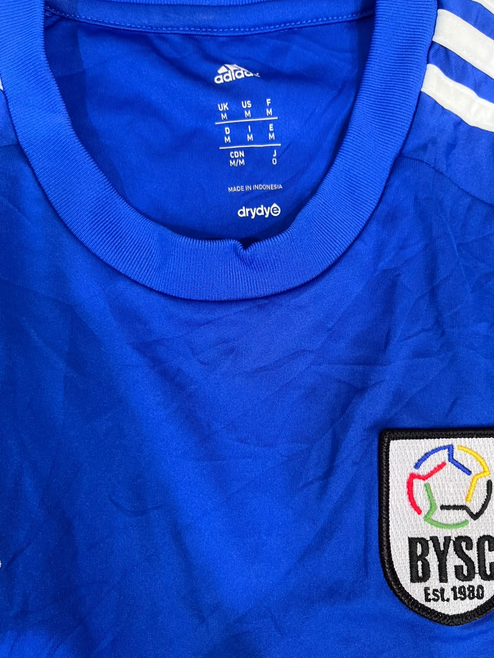 Close-up of a royal blue Adidas soccer jersey with white shoulder stripes and a BYSC Est. 1990 patch on the chest.