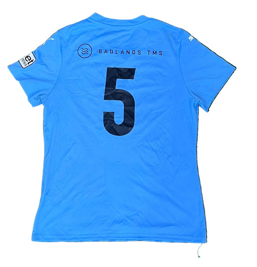 A light blue soccer jersey , showing the back side with a large black number 5 and 'BADLANDS TMS' printed above it. A small sleeve patch is visible on the left sleeve.