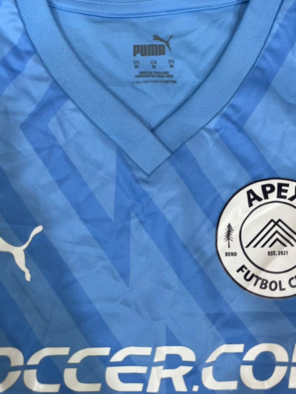 Close-up of a light blue Puma soccer jersey with a V-neck collar, showing the Puma logo on the chest and inside the collar, and part of the APEZENDO FUTSOCLUB BEND brand crest.