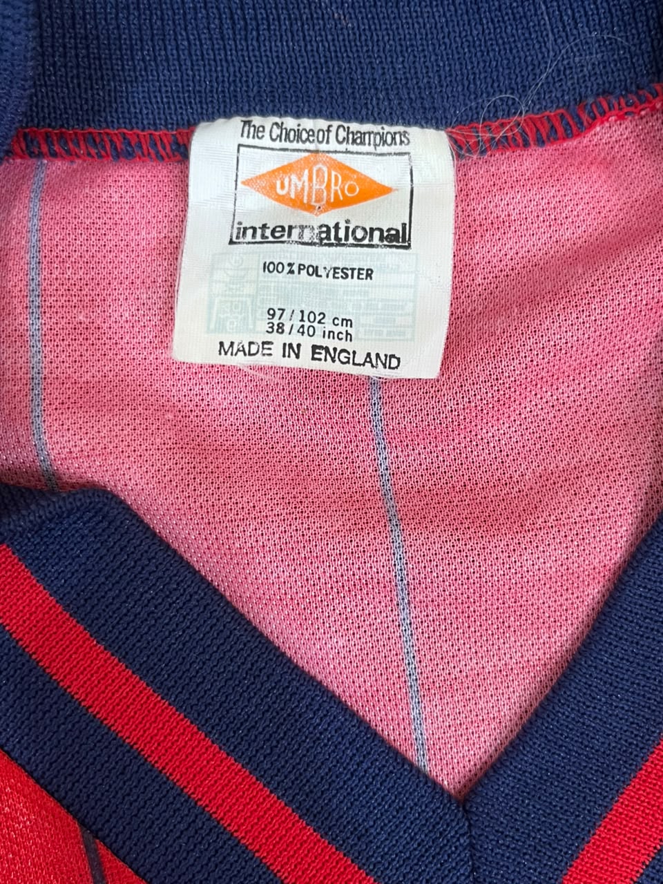 Close-up of a vintage UMBRO International pullover sweater showing the inner collar with a white size tag, pink interior lining, and navy and red striped knit fabric.