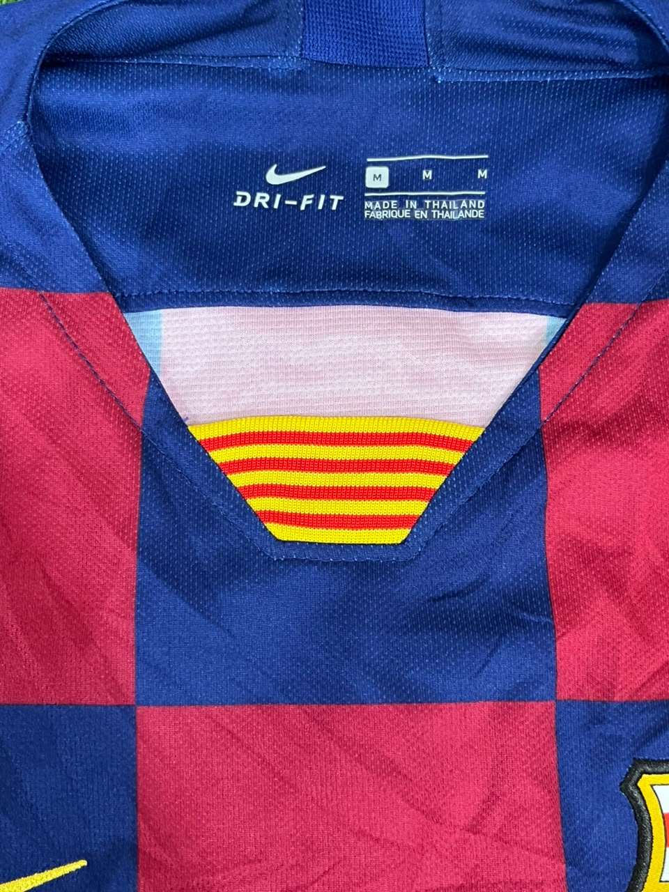 Close-up of a FC Barcelona soccer jersey showing the blue Nike Dri-FIT collar with size markings, the red and blue checkered pattern, and the yellow and red striped V-neck detail.