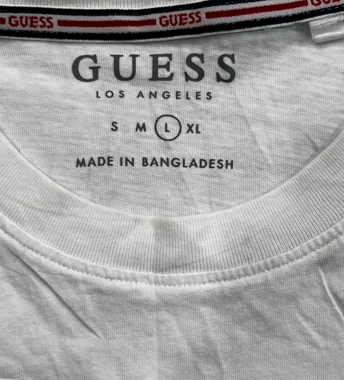 Close-up of the inside collar of a light gray heather Guess t-shirt, showing the printed GUESS LOS ANGELES branding, size information (S M L XL), and 'MADE IN BANGLADESH' on the neck label.