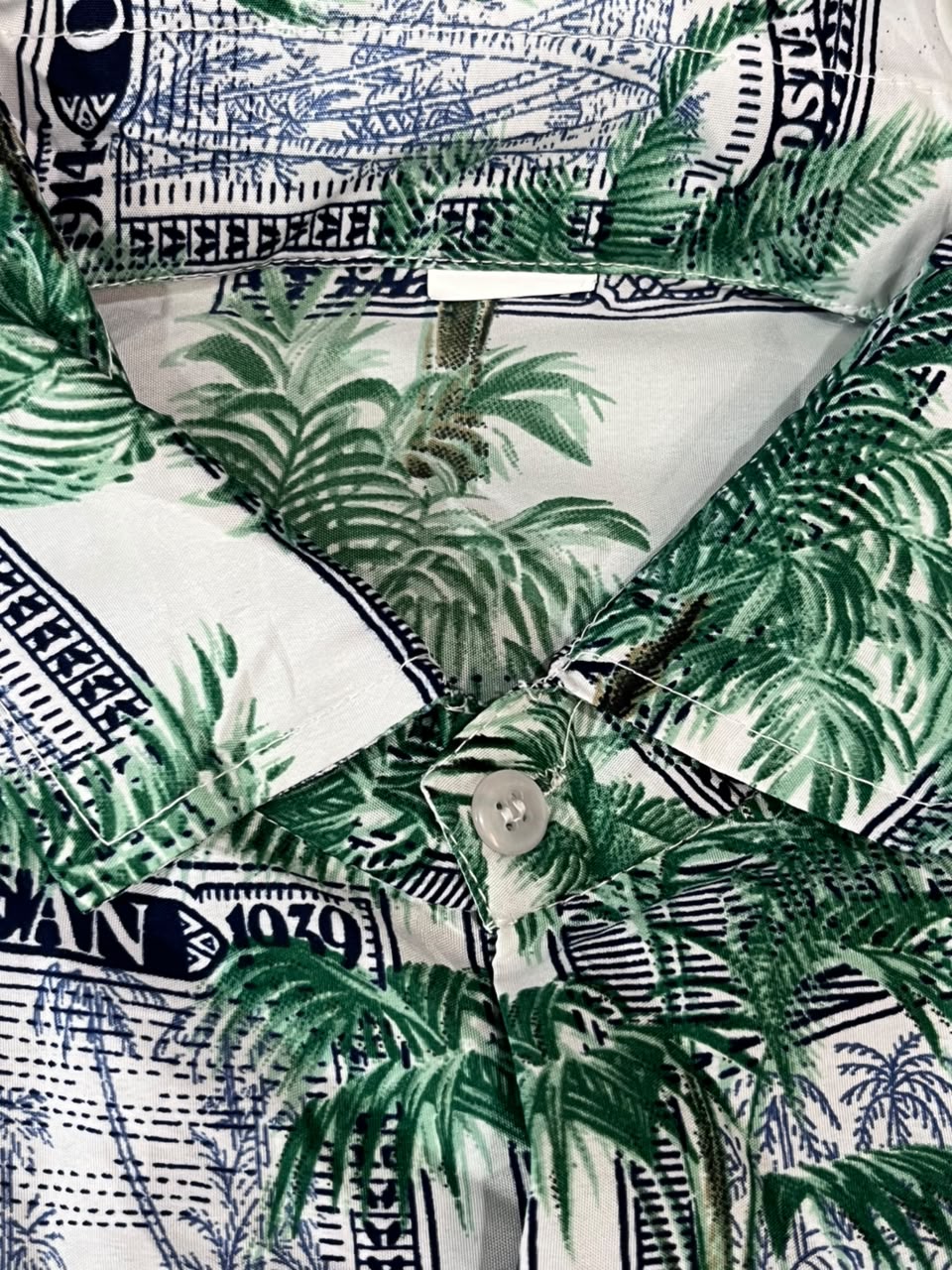 Close-up of a button-front shirt with a white base and an allover tropical palm print over a blue and black illustrated background pattern.