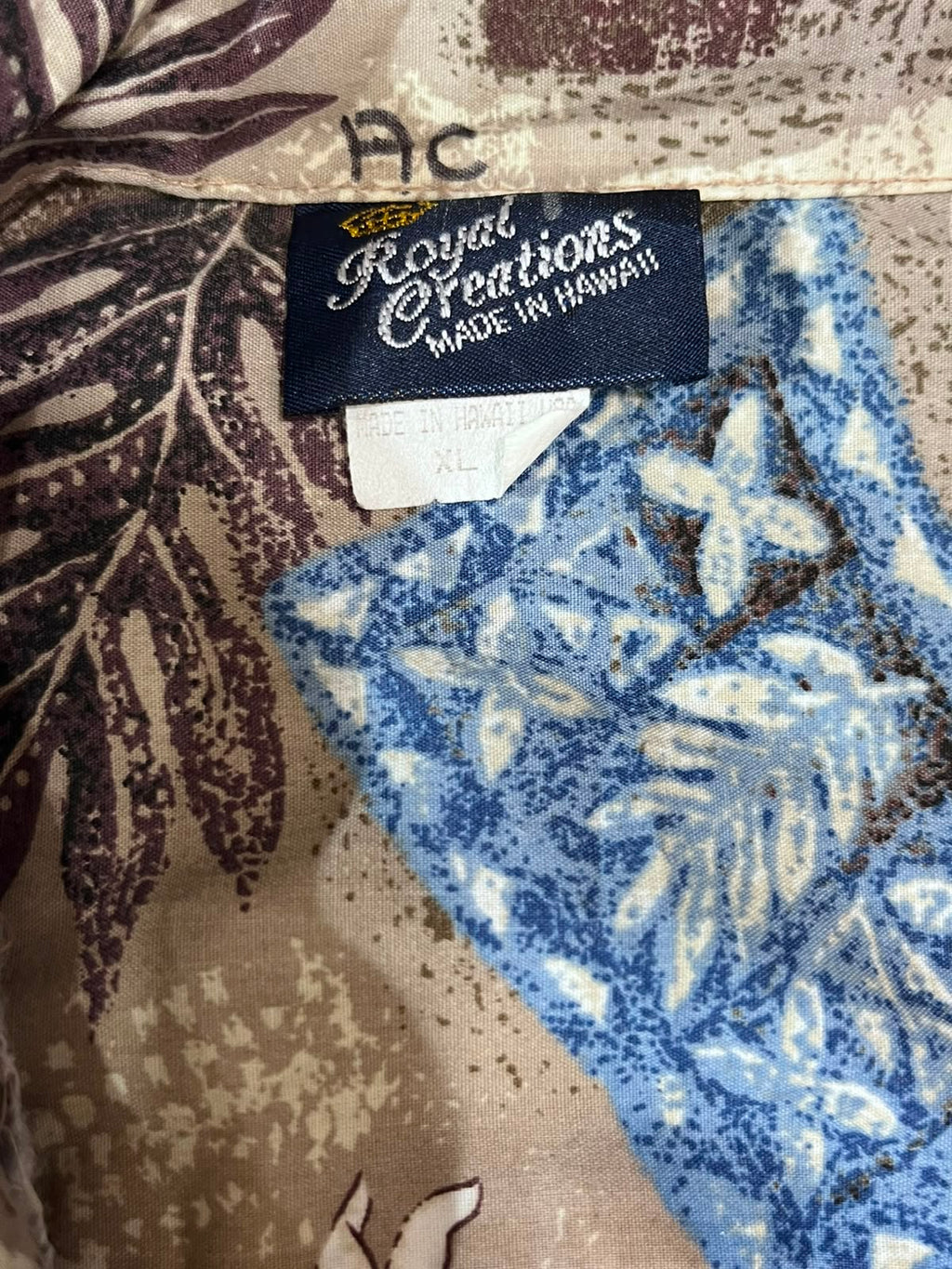 Close-up of a patterned garment label reading Royal Creations Made in Hawaii, with a blue floral print section and a beige/tan leaf-patterned fabric background.