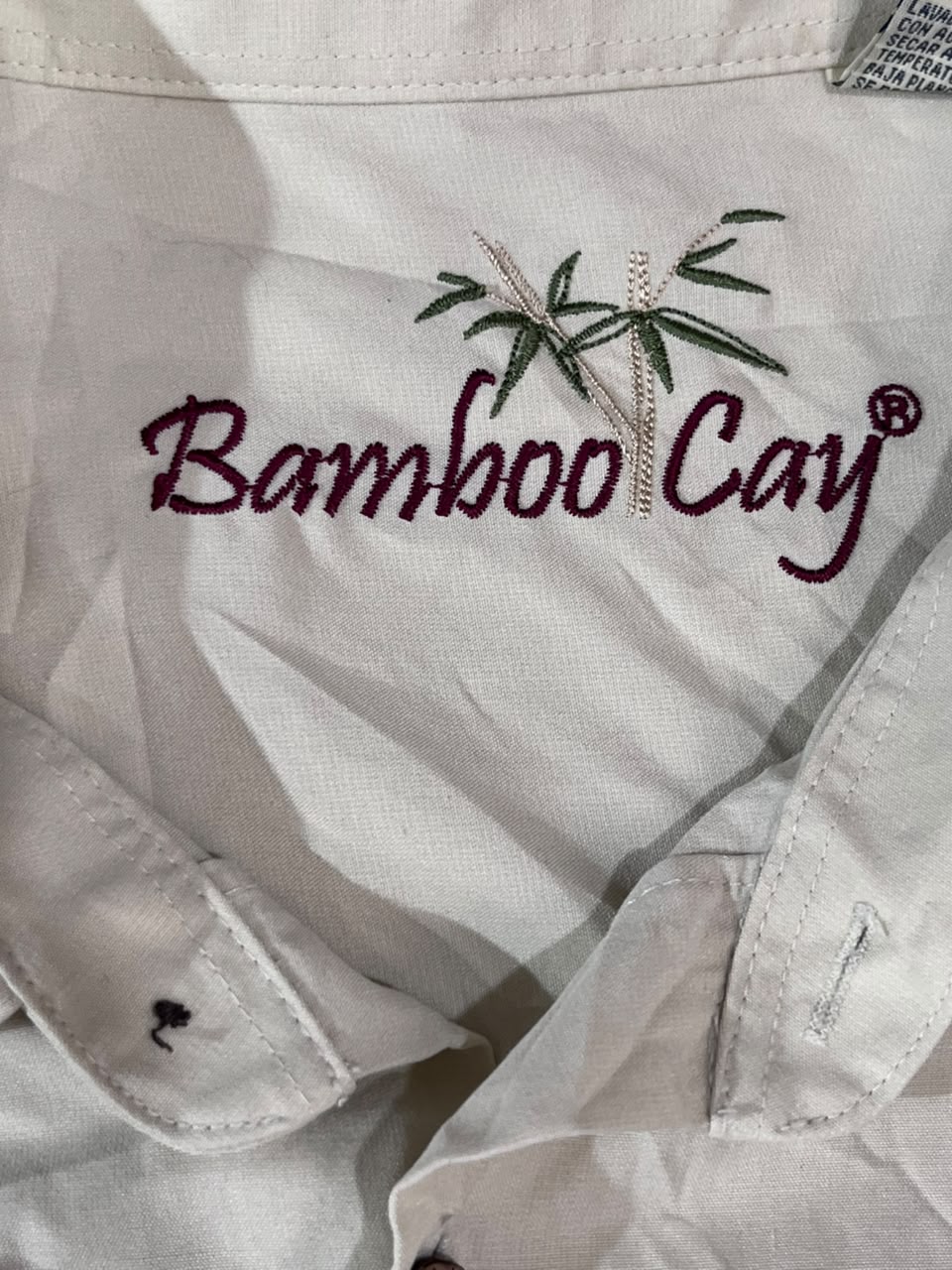 Close-up of a white or off-white fabric garment with embroidered text reading 'Bamboo Cay' in burgundy, plus a green bamboo plant motif above it.
