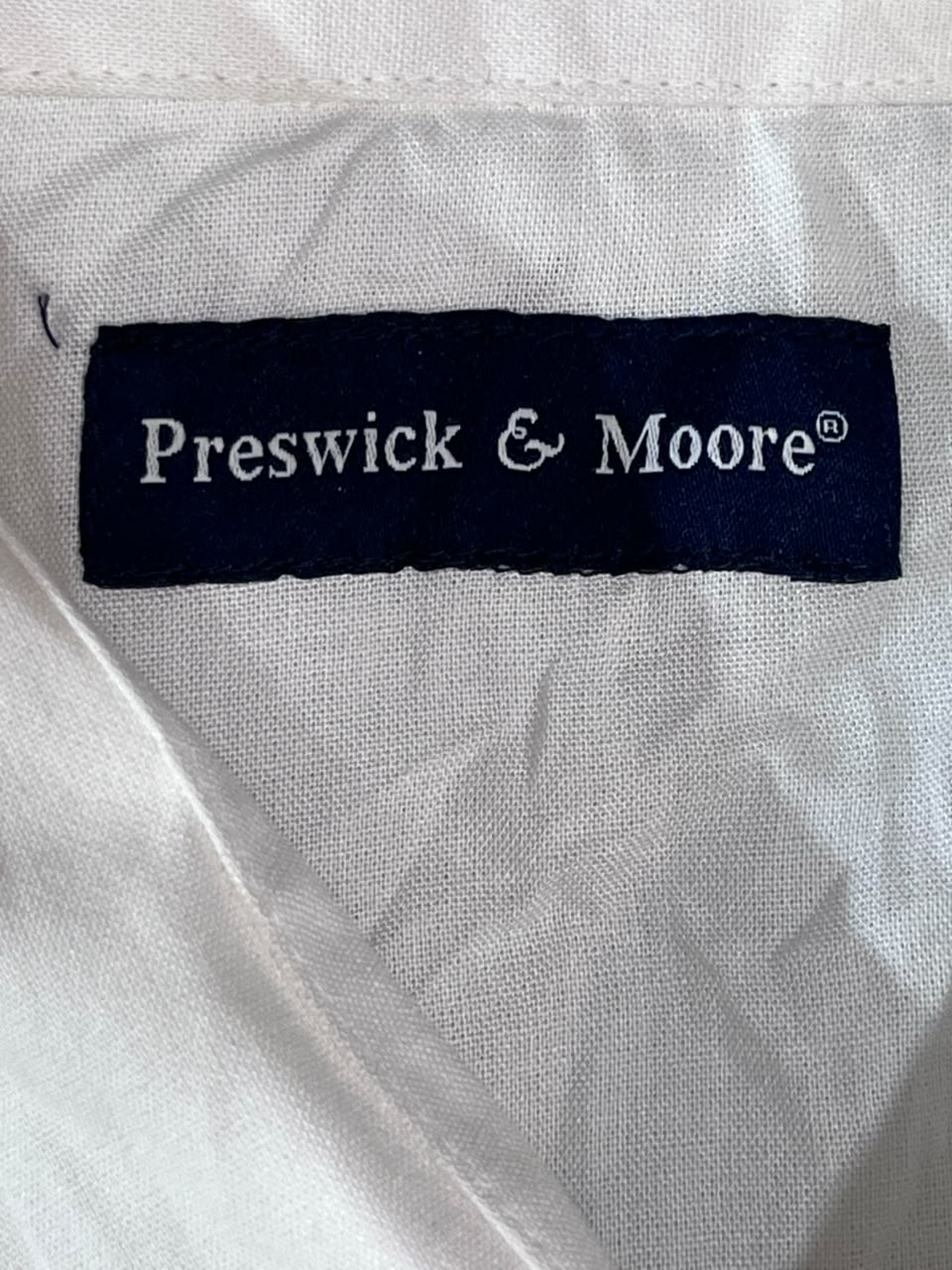 Close-up of a white fabric garment with a dark navy Preswick & Moore brand label sewn inside the collar.
