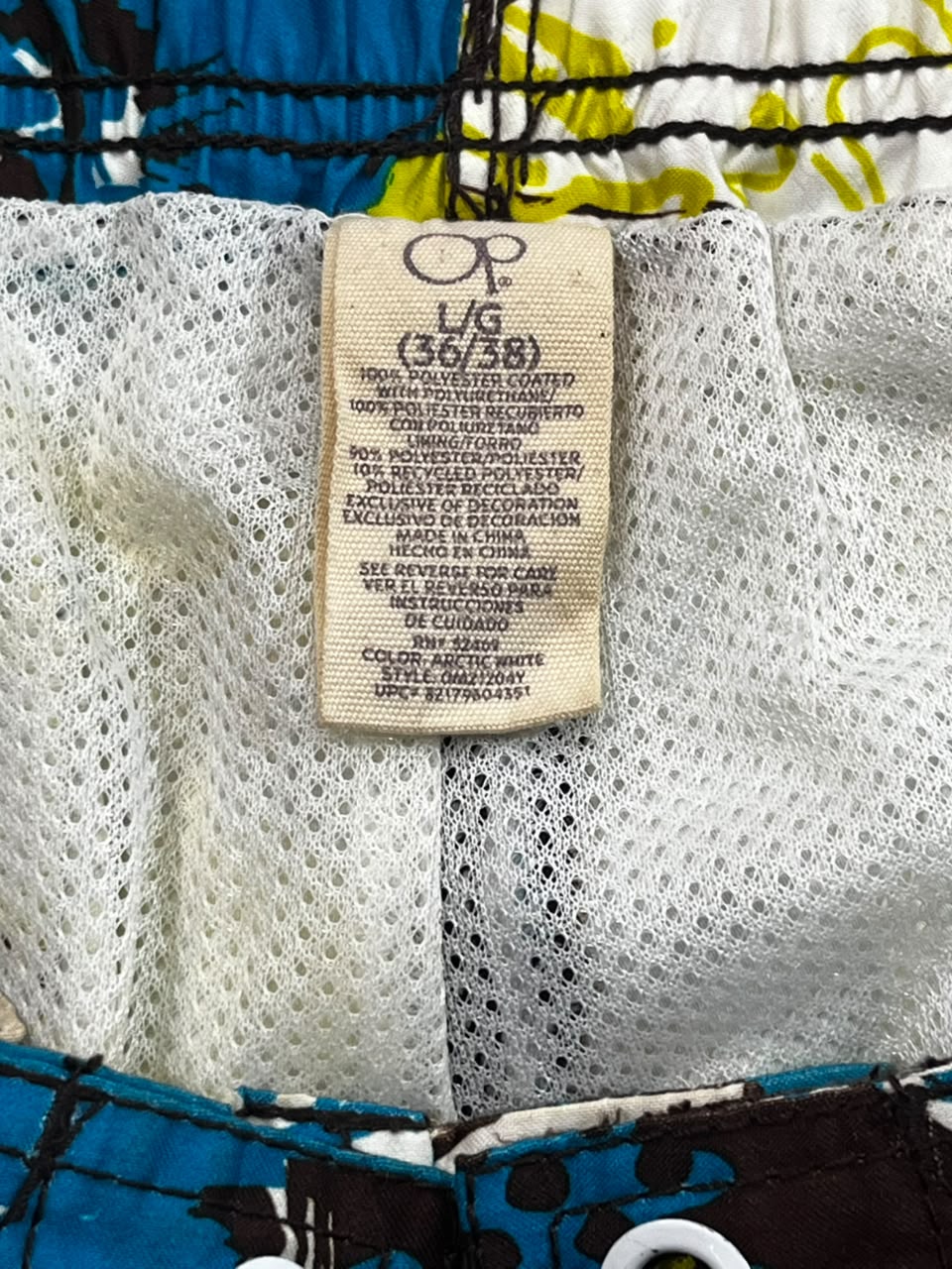 Close-up of the interior waistband of a children's swimsuit or shorts, showing a beige size tag labeled 'OP L/G (36/38)' sewn onto white mesh fabric, with blue and black patterned trim visible around it.