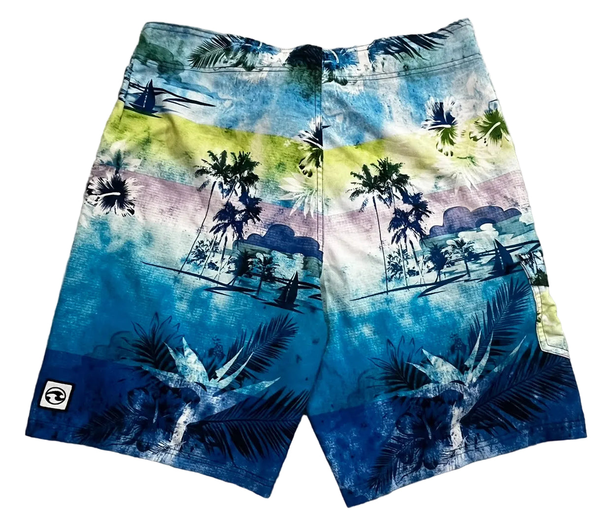A pair of men's board shorts featuring a vibrant tropical beach print with palm trees, sailboats, and a sunset gradient. The shorts are showing a blue, yellow, and pink watercolor wash pattern with dark blue accents.