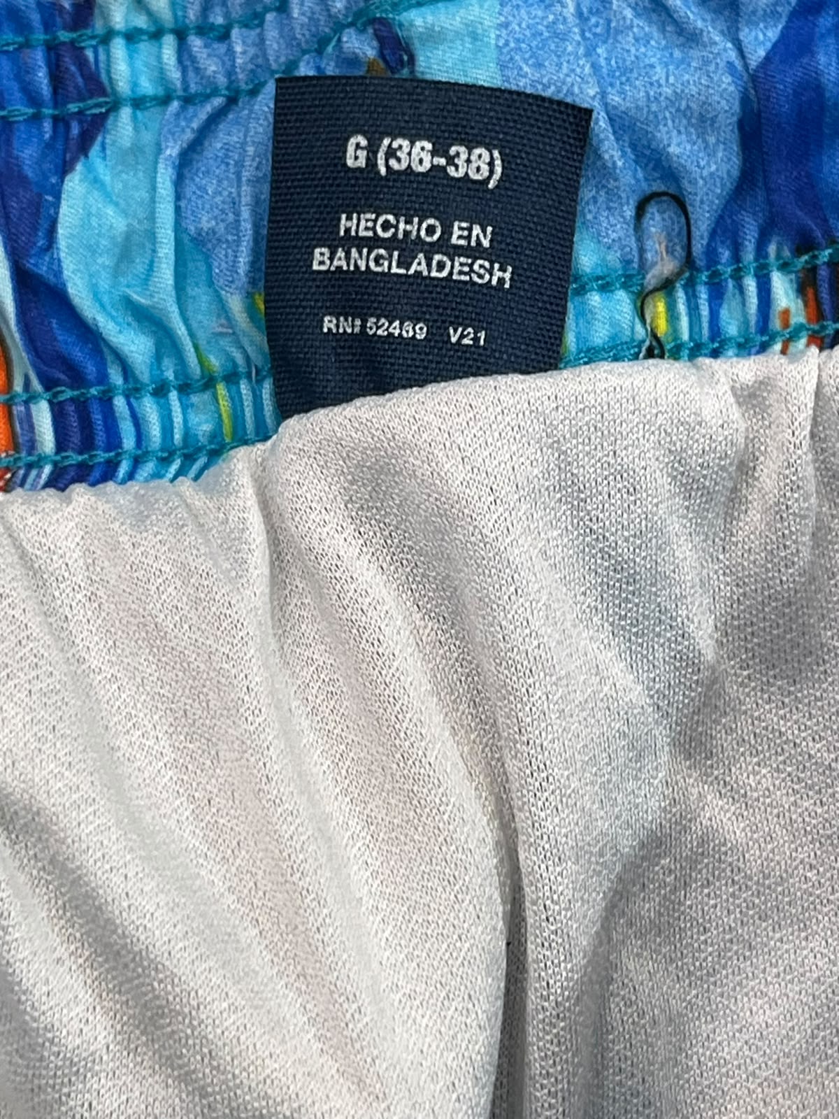 Close-up of a garment's waistband and inner label, showing a dark navy brand tag that reads 'G (38-38) HECHO EN BANGLADESH' with size and RNI details, above a light gray fabric section and blue patterned trim.