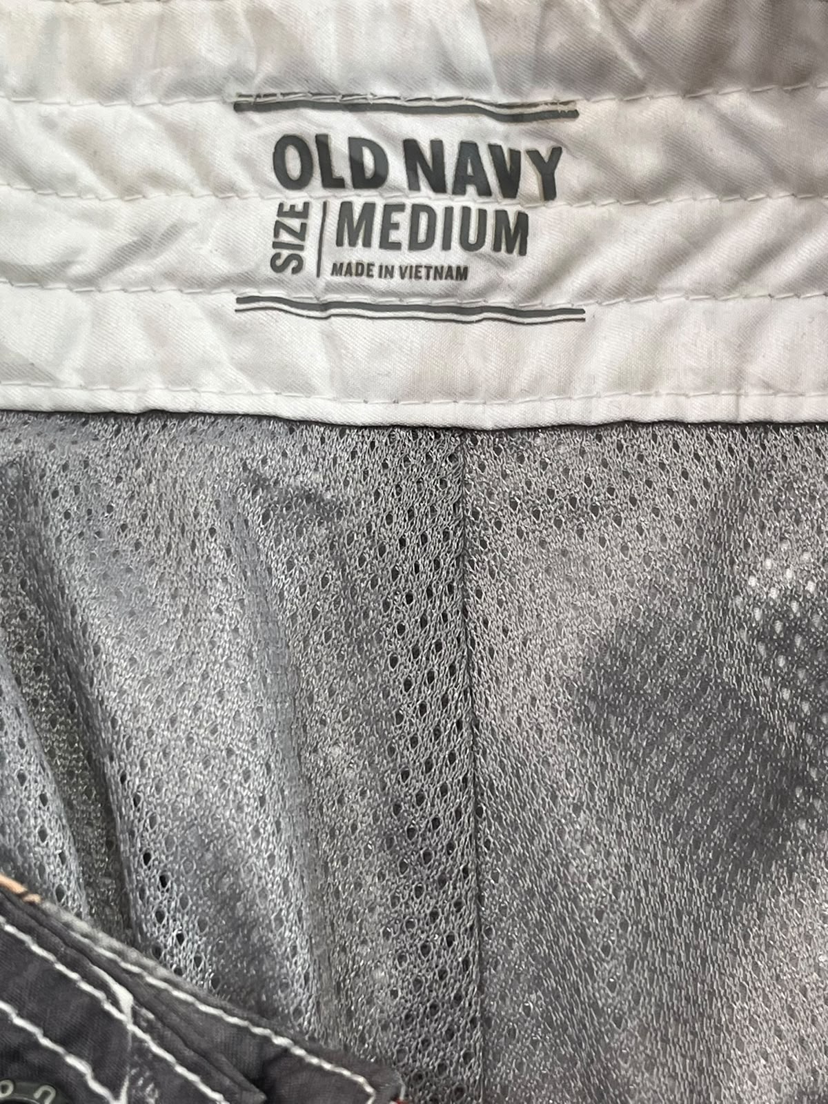 Close-up of the inside waistband of a pair of Old Navy shorts, showing the brand label 'OLD NAVY SIZE MEDIUM MADE IN VIETNAM' stitched onto a white fabric band above a gray mesh body.