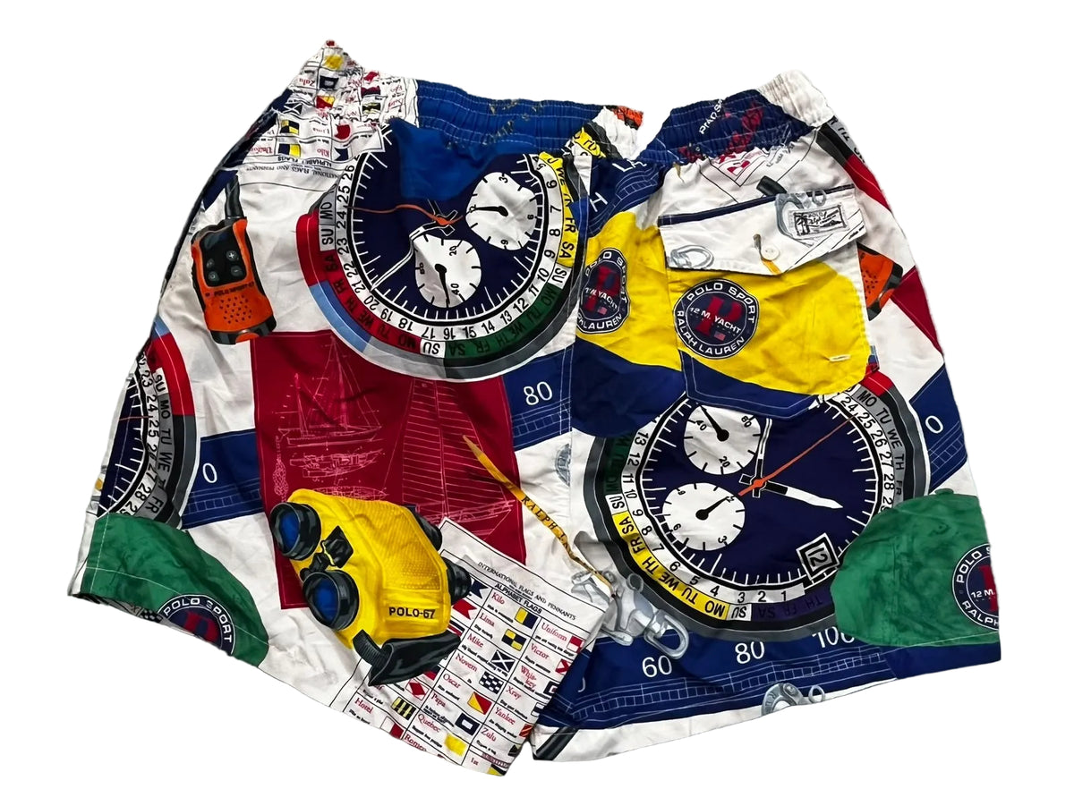 A pair of men's shorts featuring a colorful patchwork print of nautical compasses, clock faces, and maritime-themed graphics, laid flat on a gray shag rug.