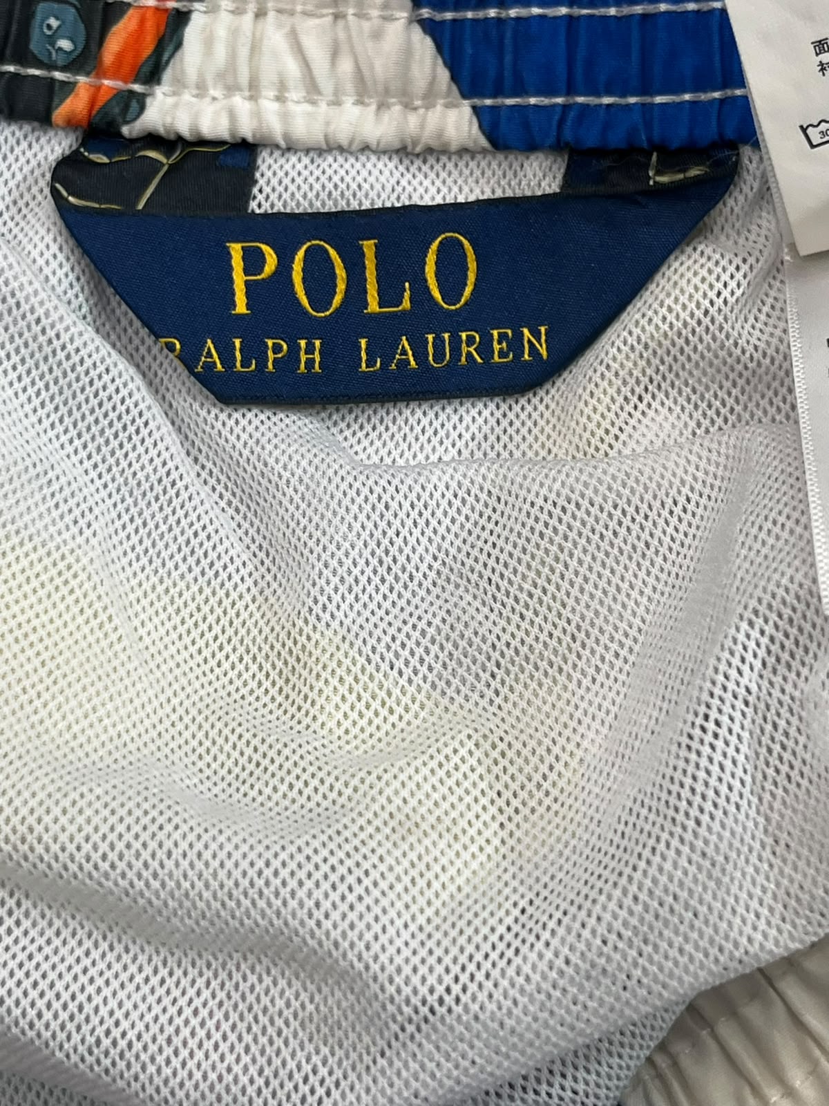 Close-up of Polo Ralph Lauren shorts showing the navy blue brand label with gold lettering, white mesh fabric, and part of the multicolor patterned waistband.