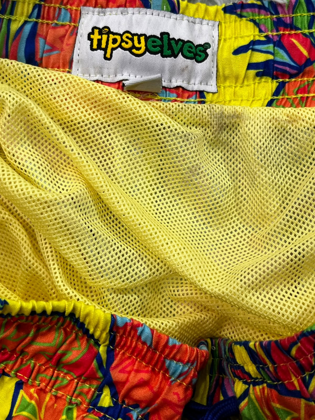 Close-up of a pair of colorful swim trunks showing a bright yellow mesh fabric, a white brand label reading 'tipsyelves', and a vibrant tropical print waistband in red, blue, green, and yellow.