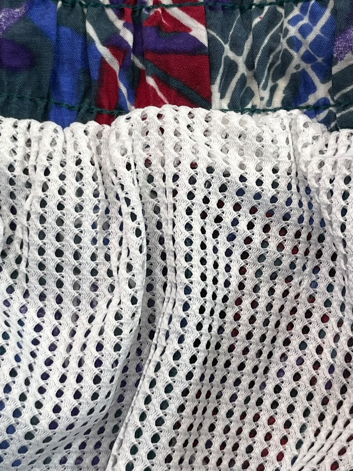 Close-up of a white open-knit mesh fabric with a gathered waistband above it, showing a colorful patterned textile in blue, red, green, and purple.