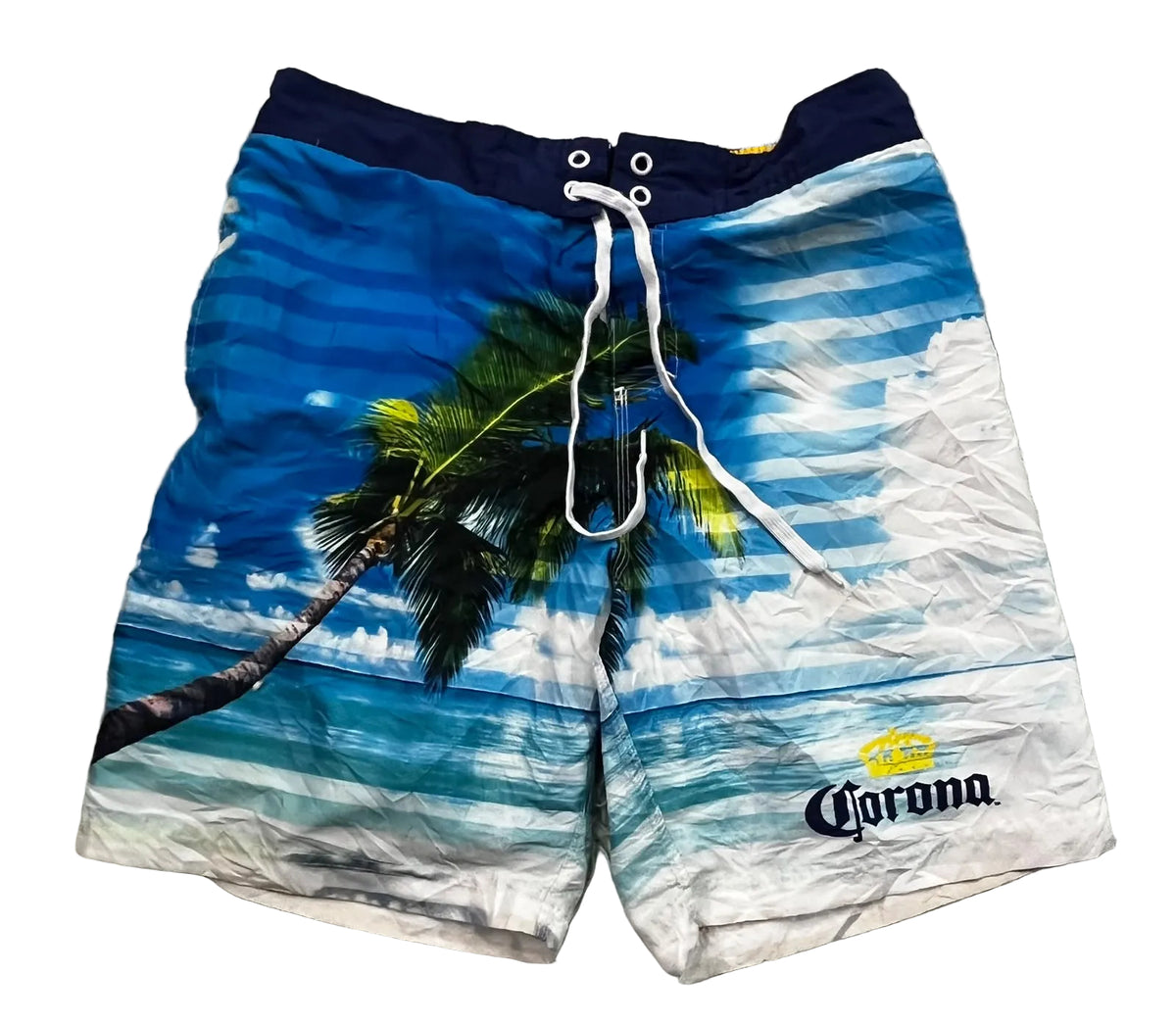 A pair of Corona brand men's swim trunks . The swim trunks feature a tropical beach print with a palm tree, blue ocean waves, and white clouds, with a navy blue waistband and white drawstrings.