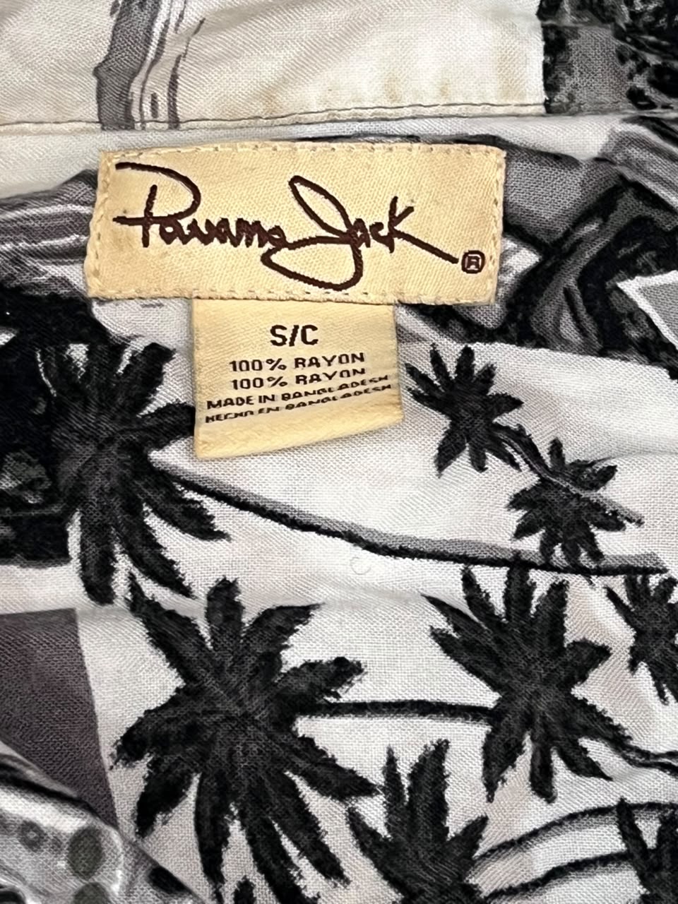 Close-up of a Panama Jack shirt fabric with a black palm tree pattern on a white background, showing the brand label and size tag.