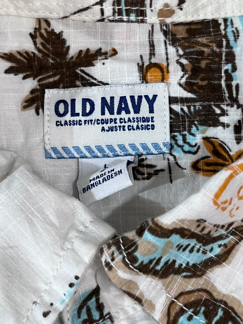 Close-up of a patterned fabric garment showing an Old Navy label and a second Bangladesh tag, with a white base and brown, black, and turquoise tropical-style print.