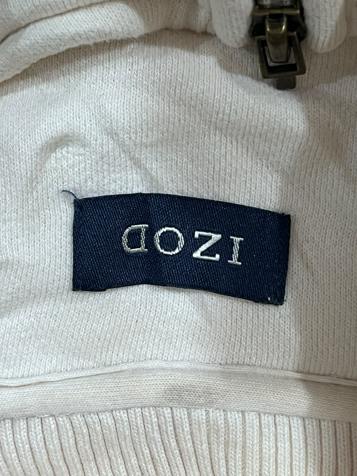 Close-up of a cream-colored ribbed knit garment with a dark navy brand label reading 'IZOD' in silver lettering.