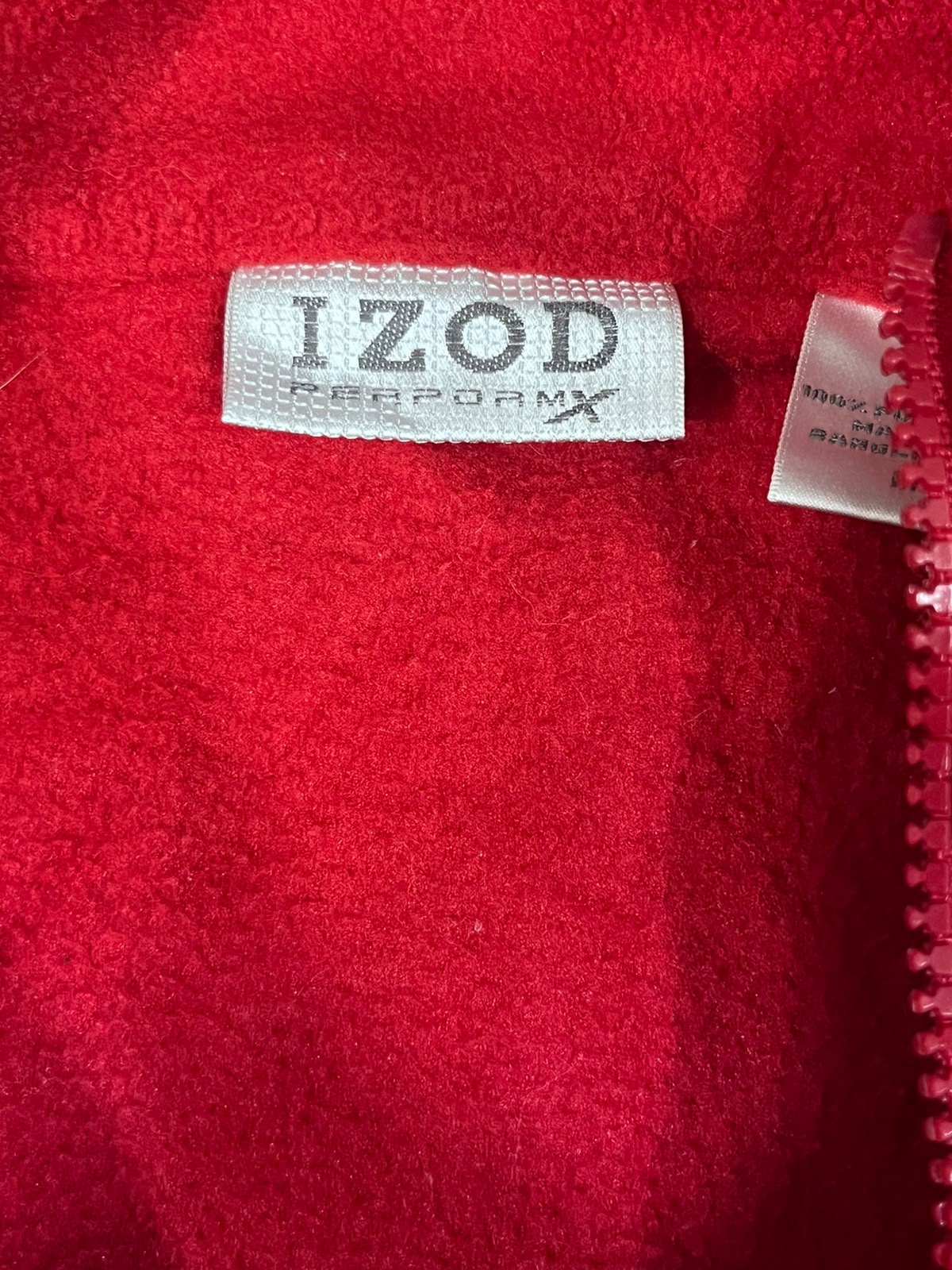 Close-up of a bright red fleece or sherpa fabric with a sewn-on label reading 'IZOD REPOAMX' and a zipper running vertically along the right side.