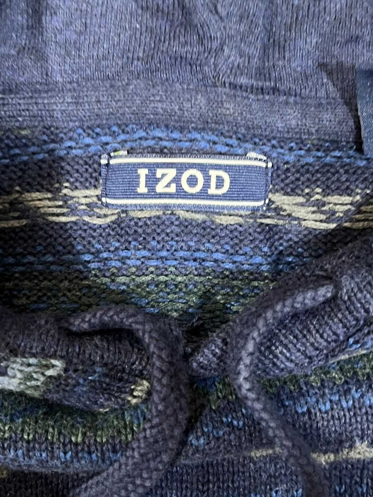 Close-up of a knit garment label reading 'IZOD' in white on a blue background, with blue, navy, green, and beige striped knit fabric visible around it.