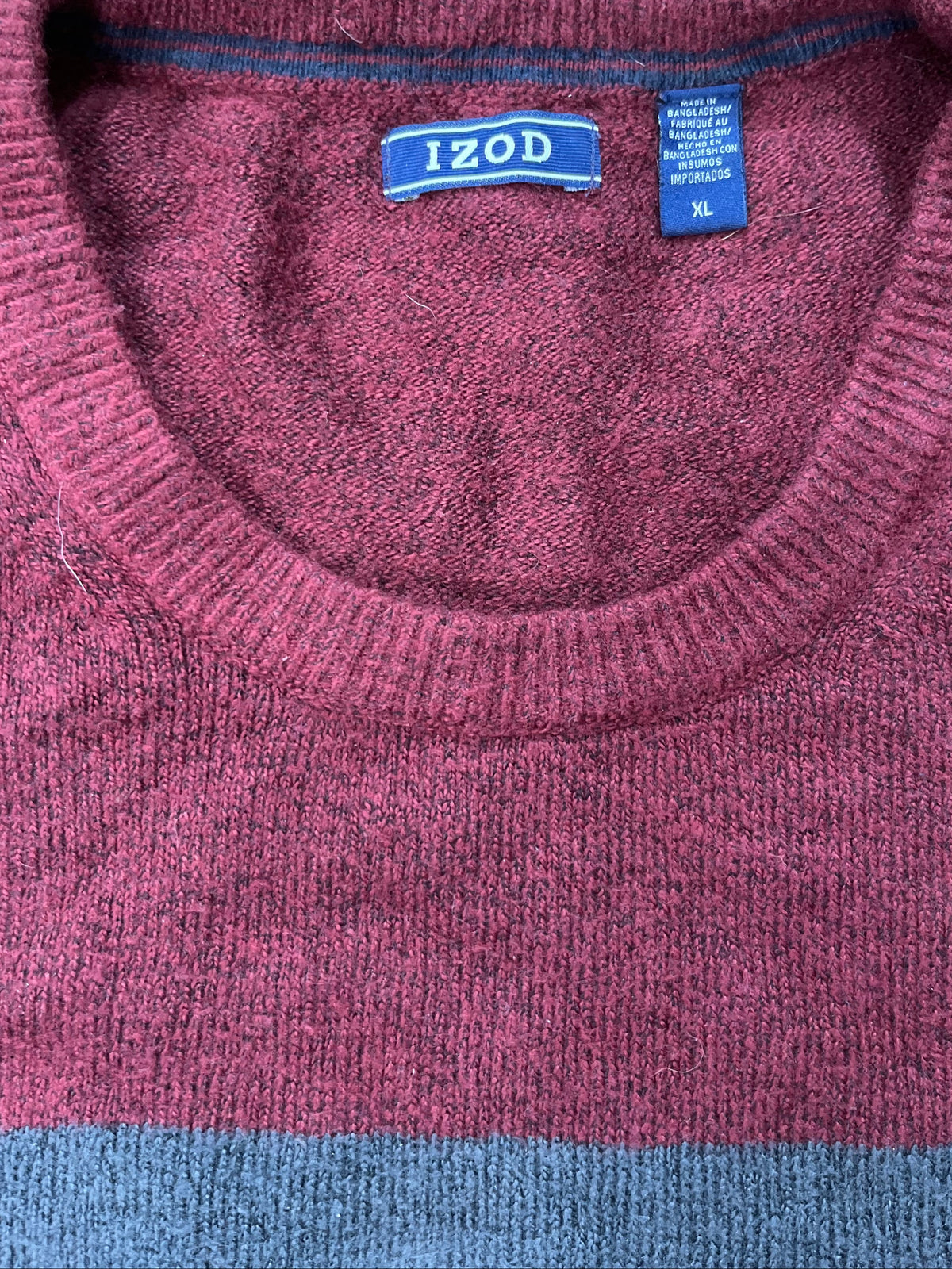 A close-up of a knit sweater by Izod, featuring a heathered burgundy body with a light blue horizontal stripe at the bottom and a blue neck label.